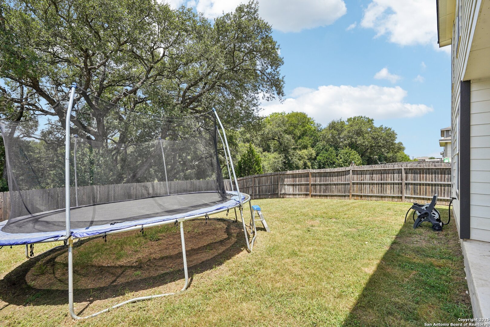 Fenced backyard with a large trampoline, mature trees, and a chair beside the house.