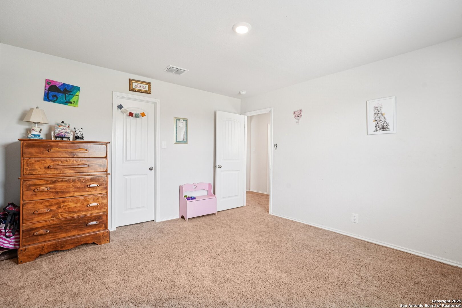 Furnished bedroom with beige carpet, a wooden dresser, white doors, and a small pink bench.