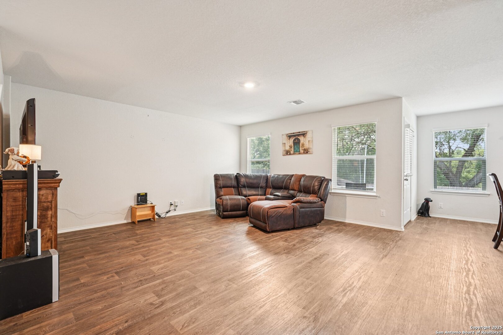 Living room with a brown leather sectional, wood-look floors, and windows overlooking trees.
