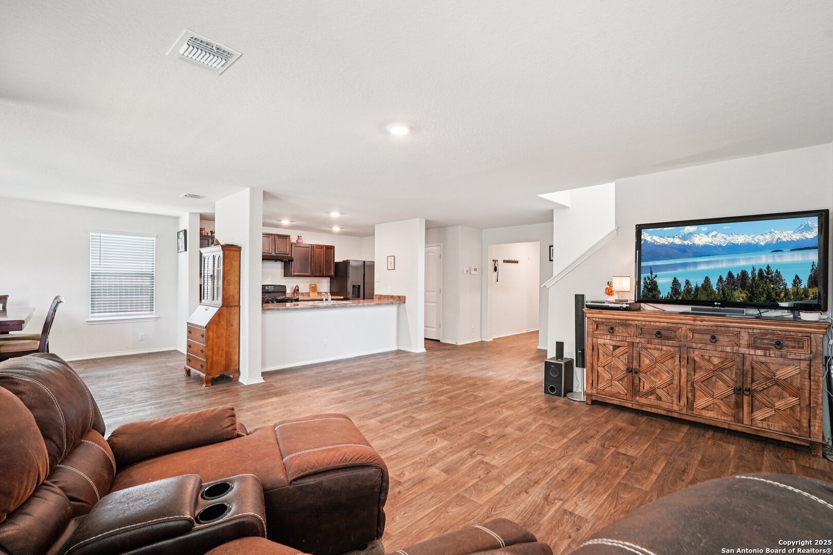 Open-plan living room with a brown leather sectional, wood-look floors, and a kitchen breakfast bar.