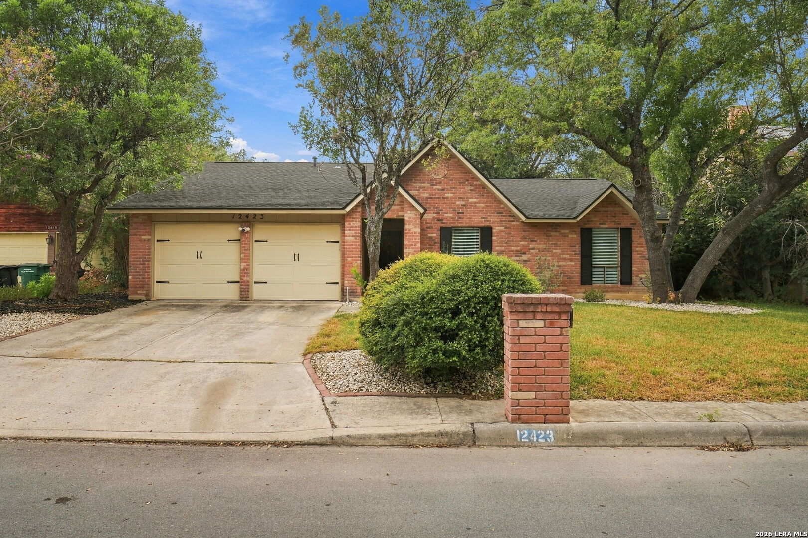 Red brick single-story home with two cream garage doors, mature trees, and a front lawn.