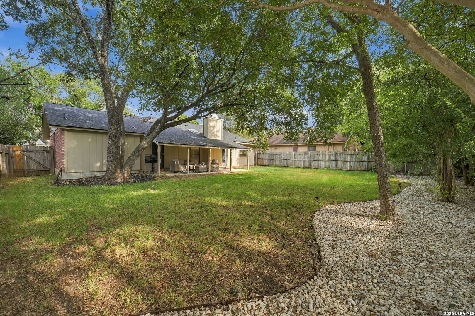 Fenced backyard with mature shade trees, a covered patio, and a curving white-stone path.