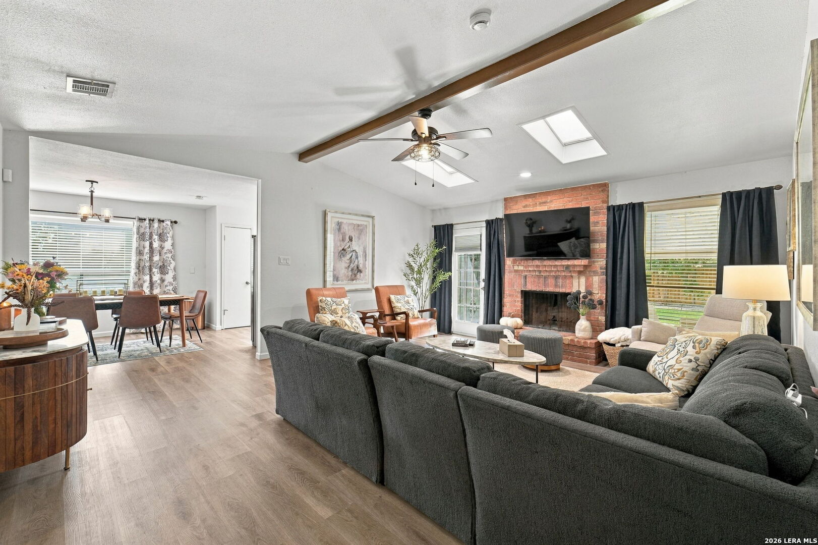 Living room with a brick fireplace, gray sectional, skylights, and an adjoining dining area.