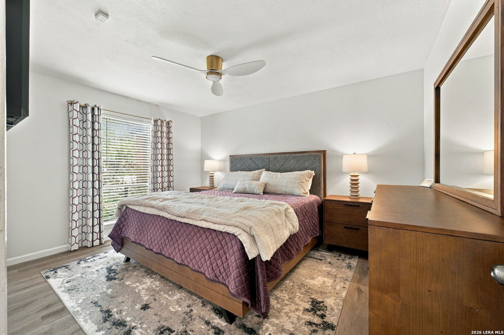 Staged bedroom with a wooden bed, plum quilt, patterned rug, and matching dresser and mirror.