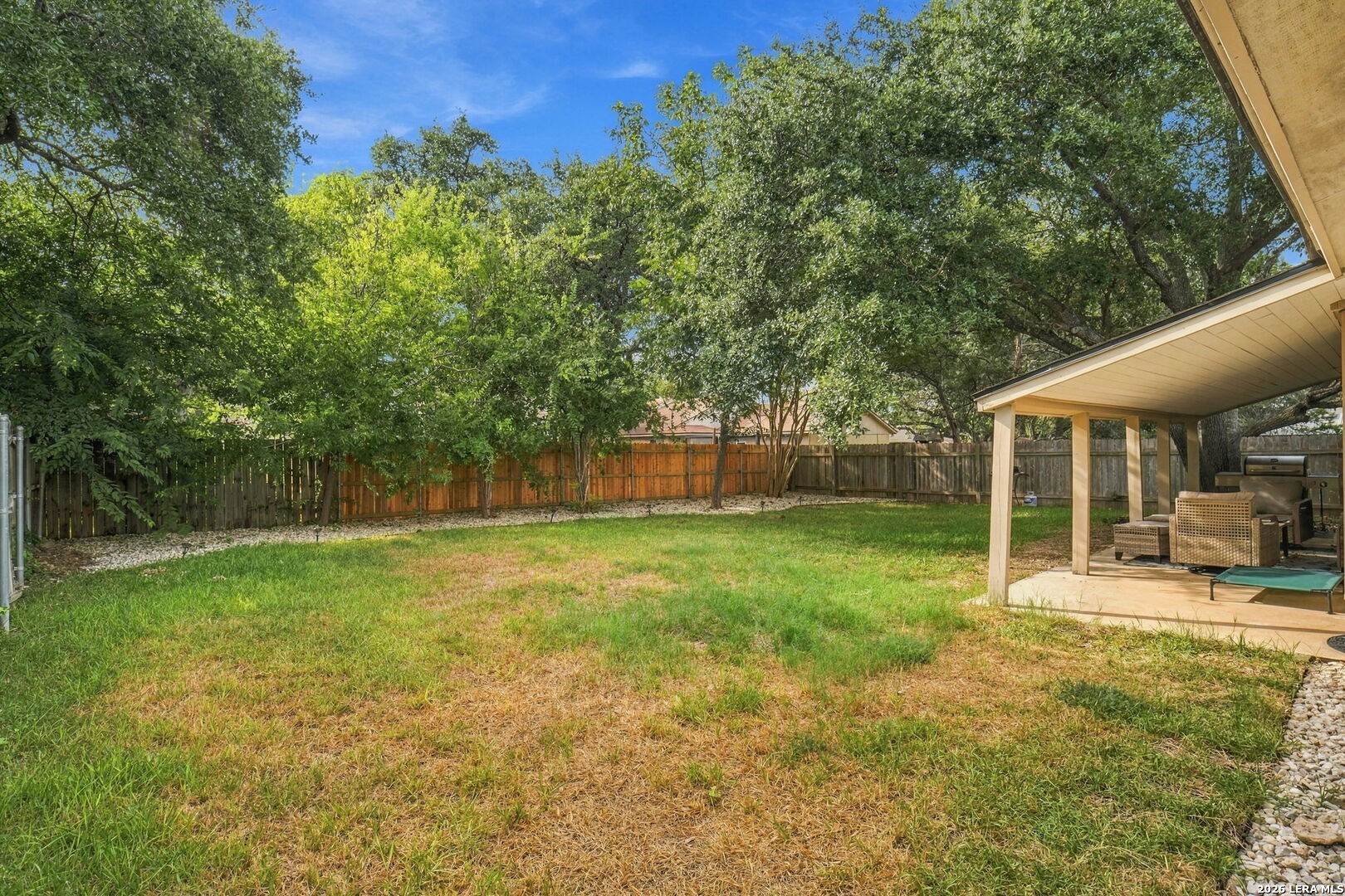 Fenced backyard with mature trees, a patchy lawn, and a covered patio with seating and a grill.