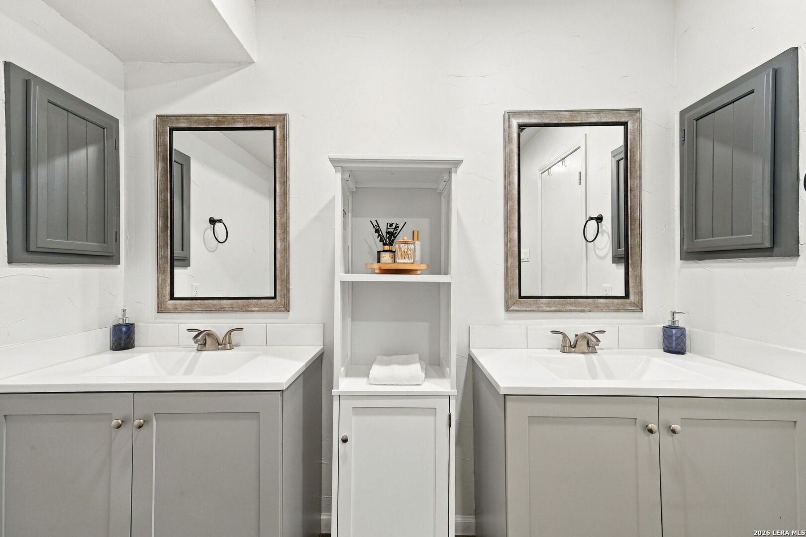 Bathroom with paired gray vanities and framed mirrors flanking central built-in shelving.