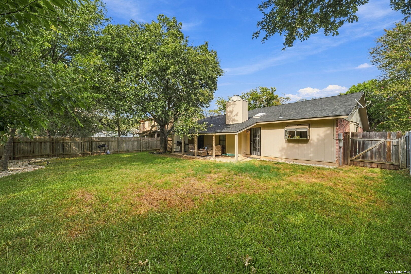 Fenced backyard with a broad lawn, mature trees, and covered patio with outdoor seating.
