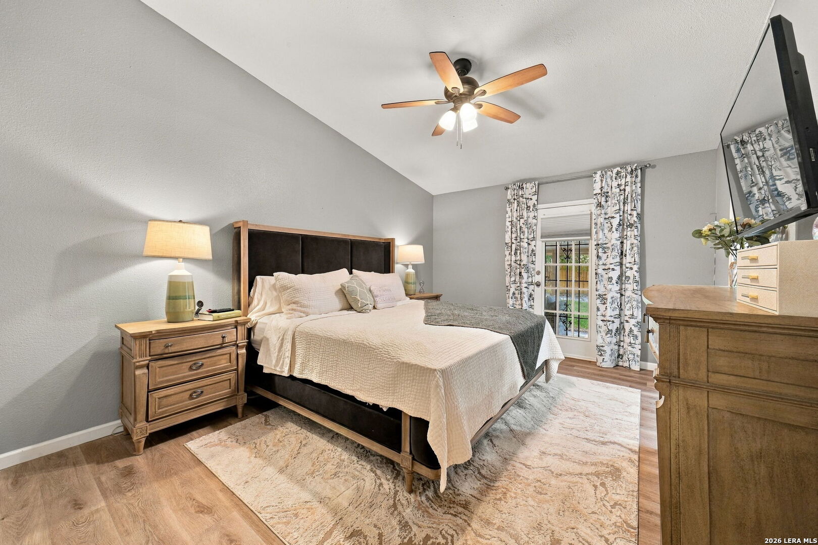 Furnished bedroom with a large bed, wood furniture, patterned rug, and glass door with floral curtains.
