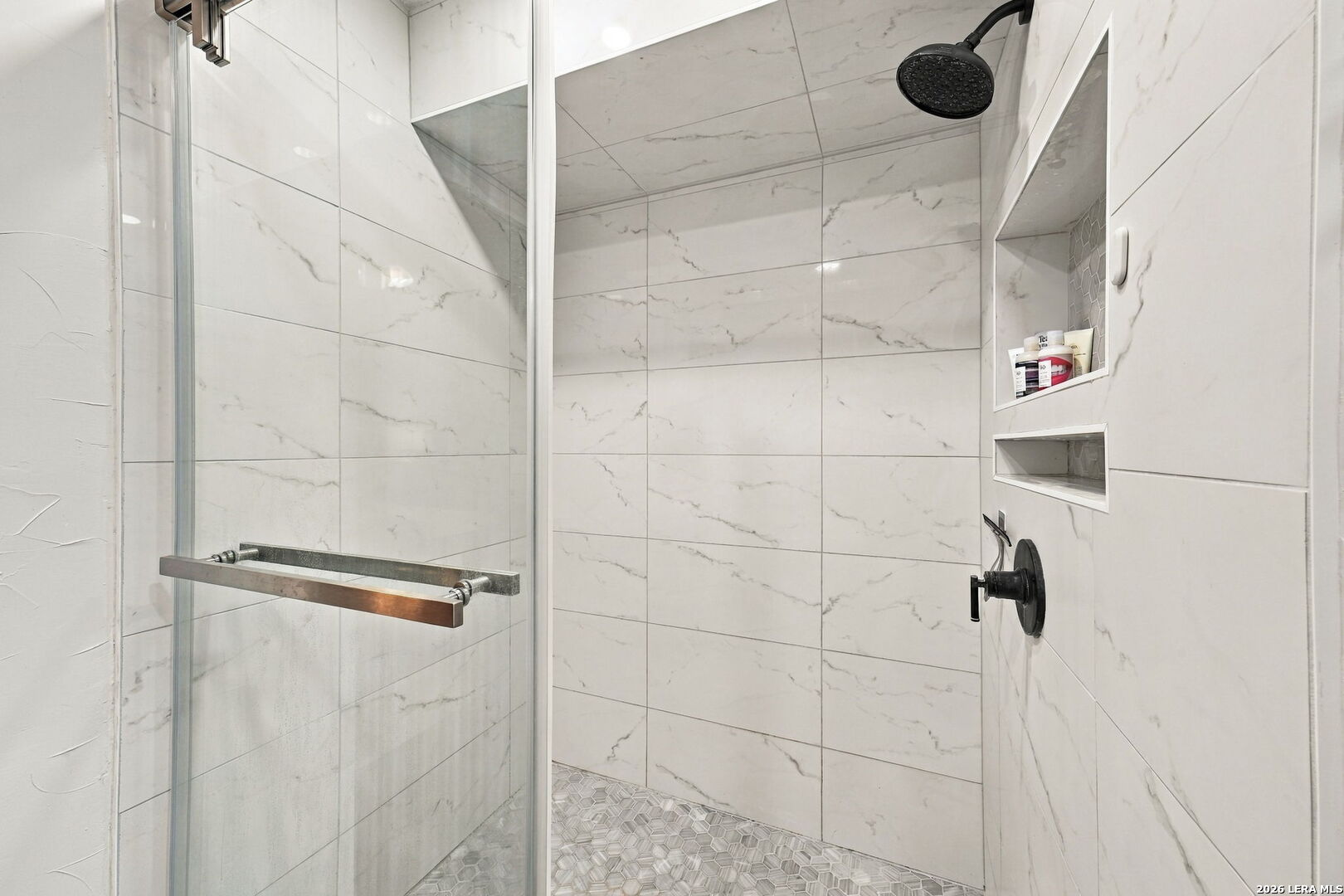 Walk-in shower with marble-look tile, black fixtures, and a gray geometric mosaic floor.