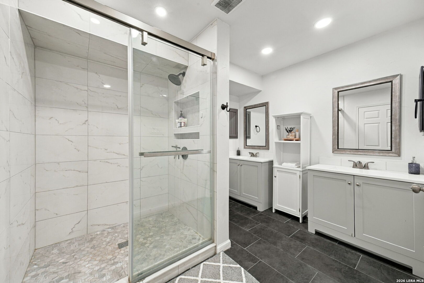 Bathroom with a glass-enclosed marble-look shower and gray double vanities.