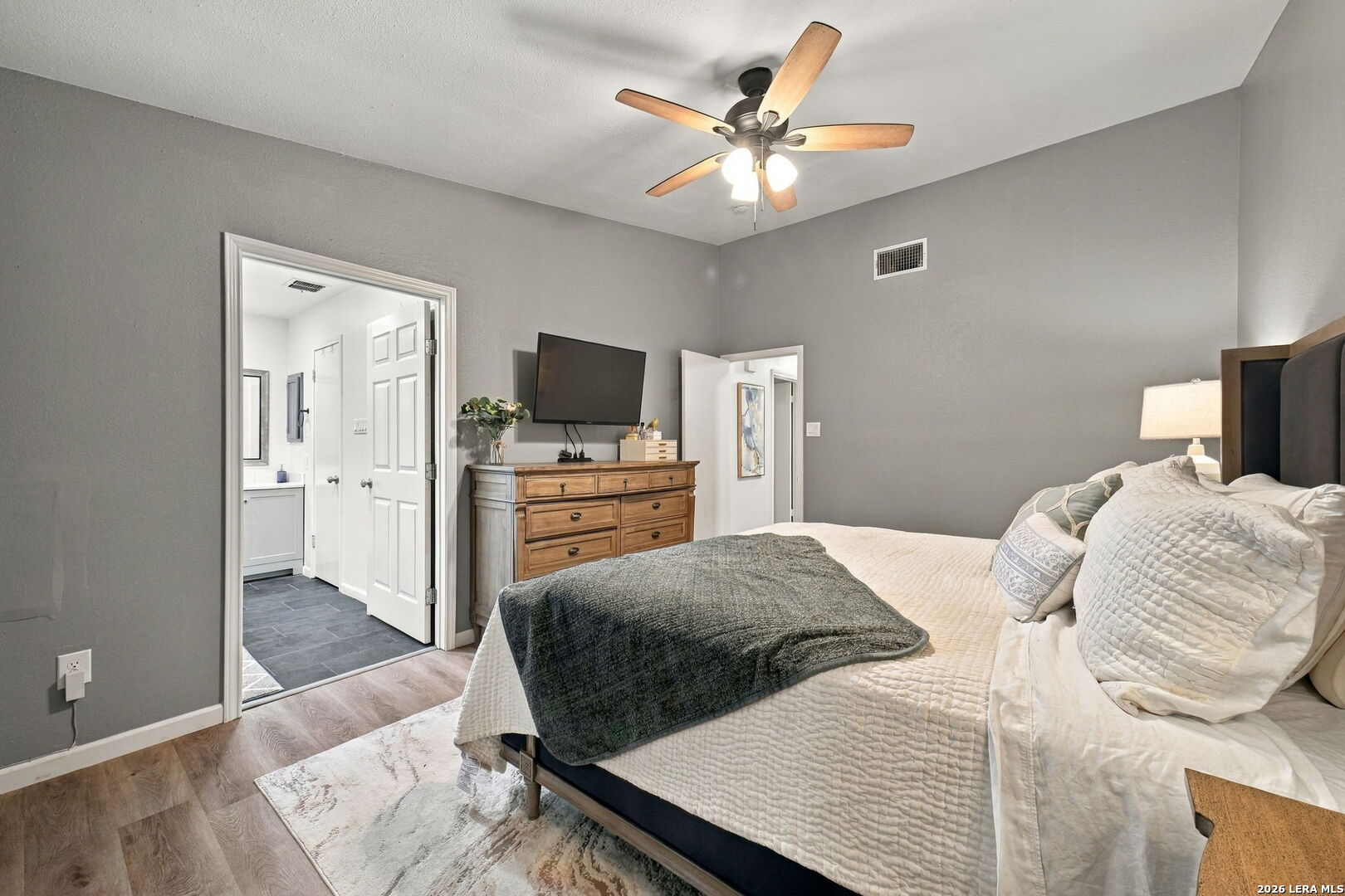 Bedroom with a large bed, gray walls, wood dresser, and ceiling fan beside an open bathroom doorway.