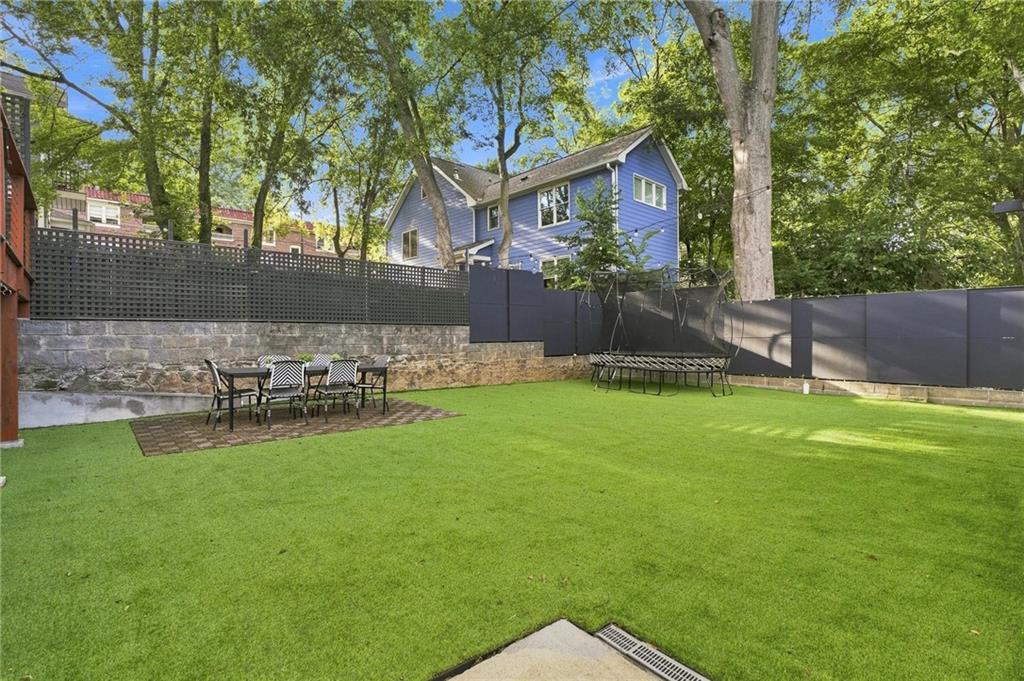 Backyard with a broad green lawn, dark fencing, stone retaining wall, and seating beneath mature trees.