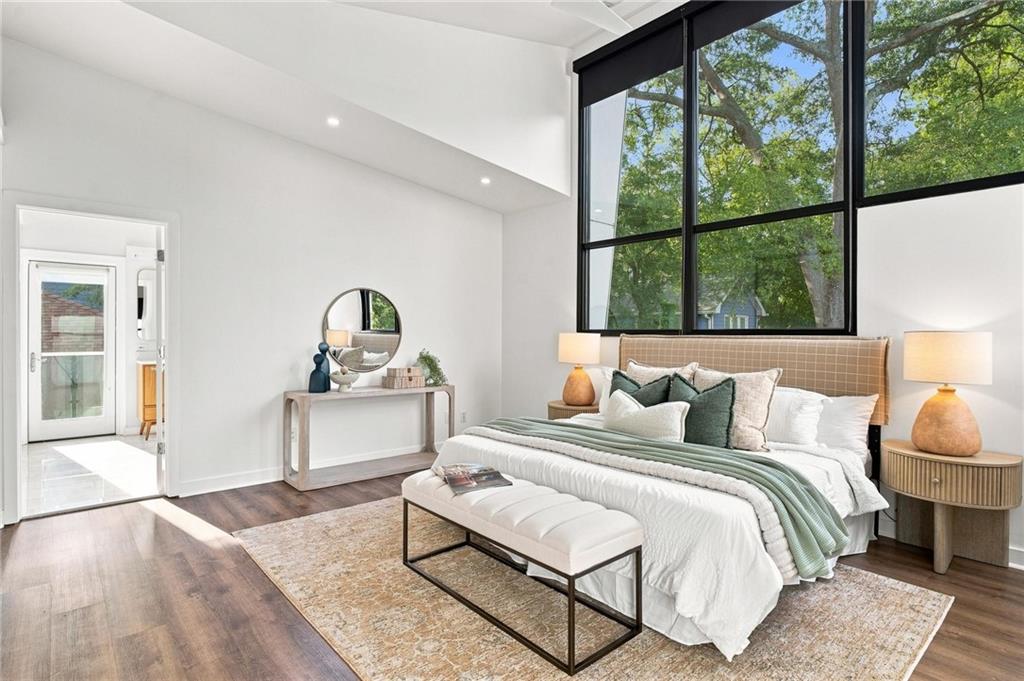 Staged bedroom with a white bed beneath tall black-framed windows overlooking leafy trees.