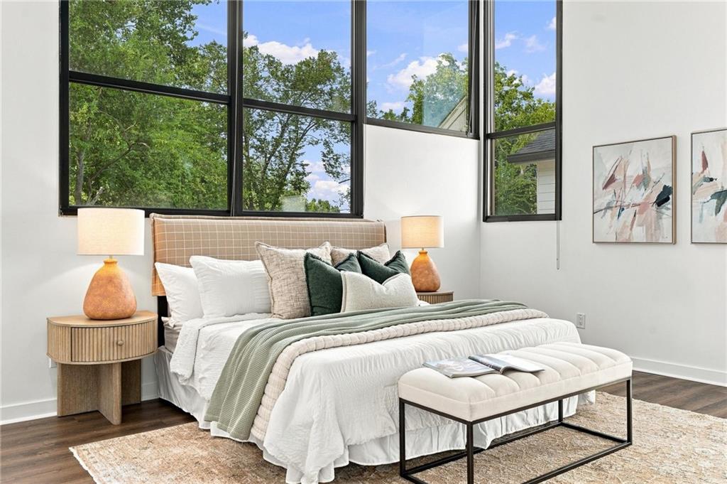 Staged bedroom with a white bed, upholstered bench, and tall windows overlooking leafy trees.