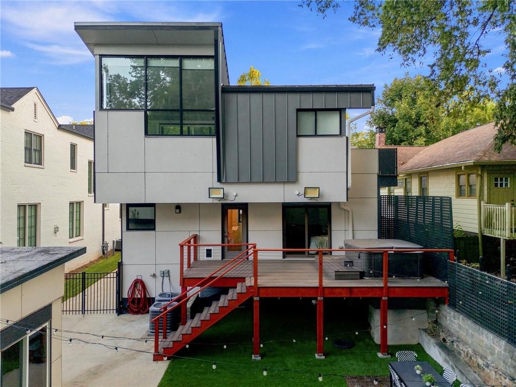 Modern home’s rear exterior with a raised deck, red railings, and stairs overlooking a small lawn.