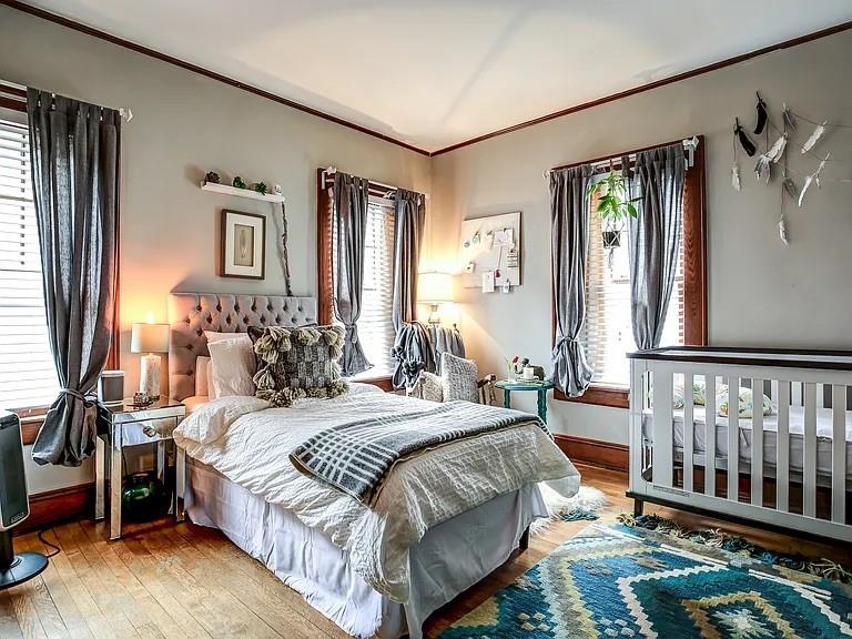 Bedroom with a tufted bed and crib beside tall curtained windows.