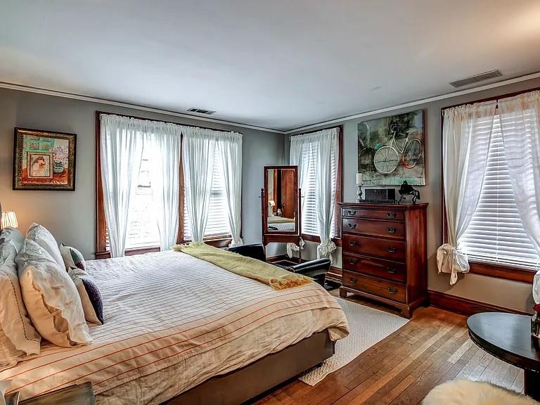 Bedroom with a large striped bed, tall curtained windows, hardwood floors, and a dark wooden dresser.