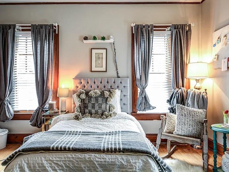 Bedroom with a tufted gray headboard, patterned bedding, and two tall curtained windows.