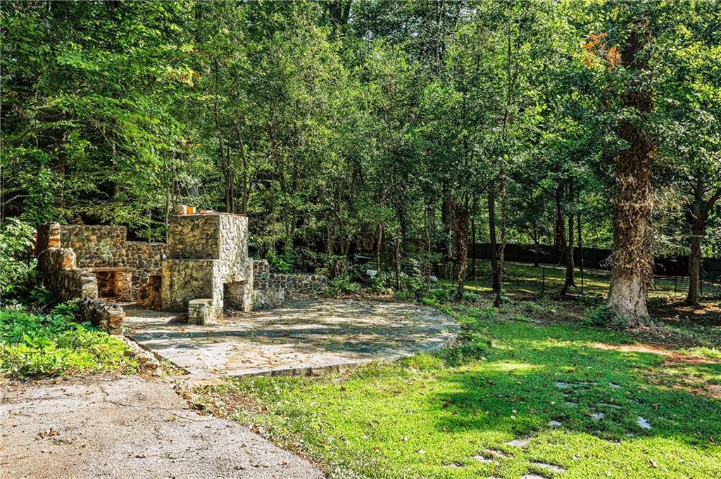 Wooded backyard with a stone patio and rustic outdoor fireplace.