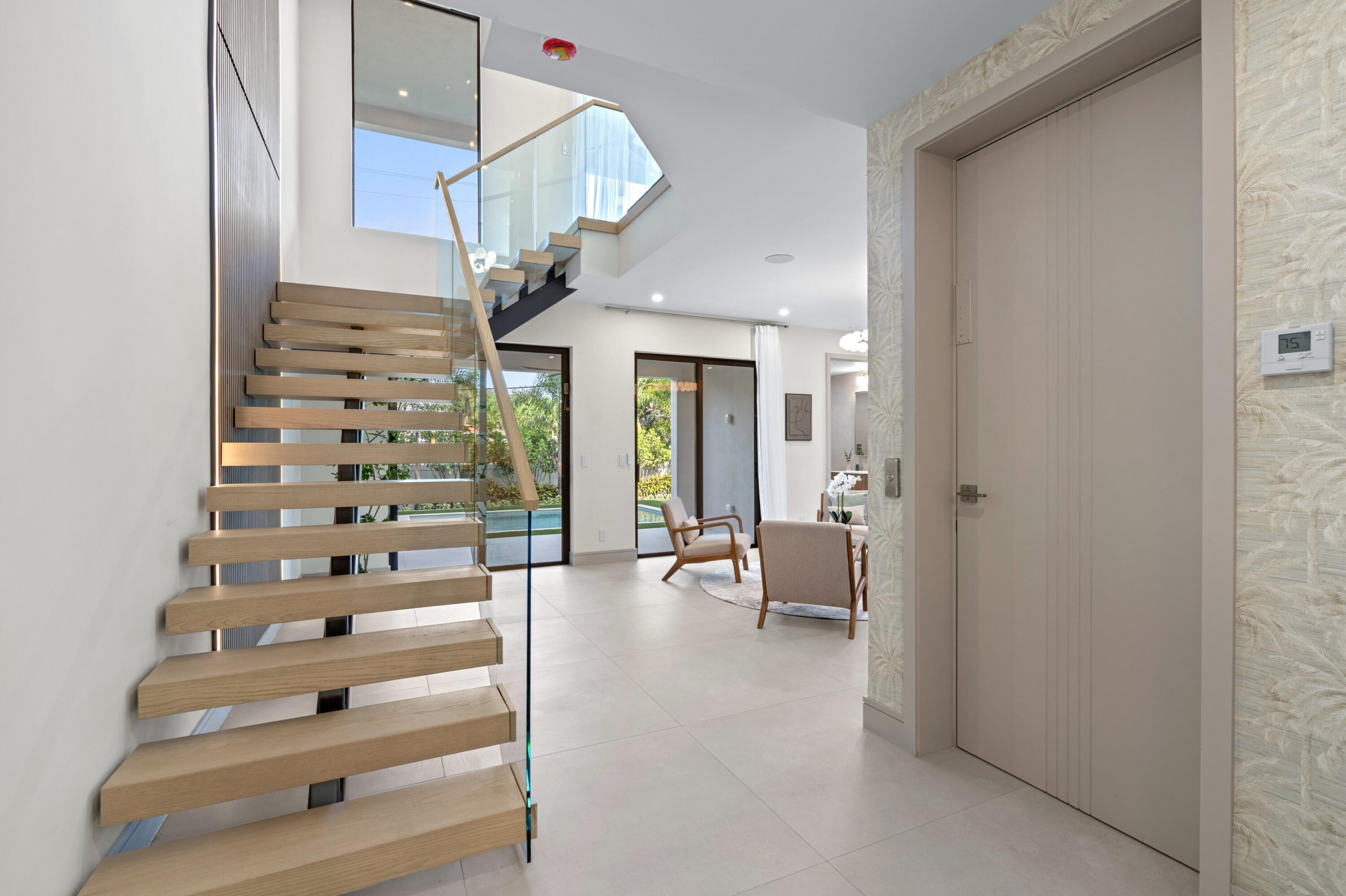 Modern entry hall with floating wood staircase, glass railing, and sitting area overlooking the garden.