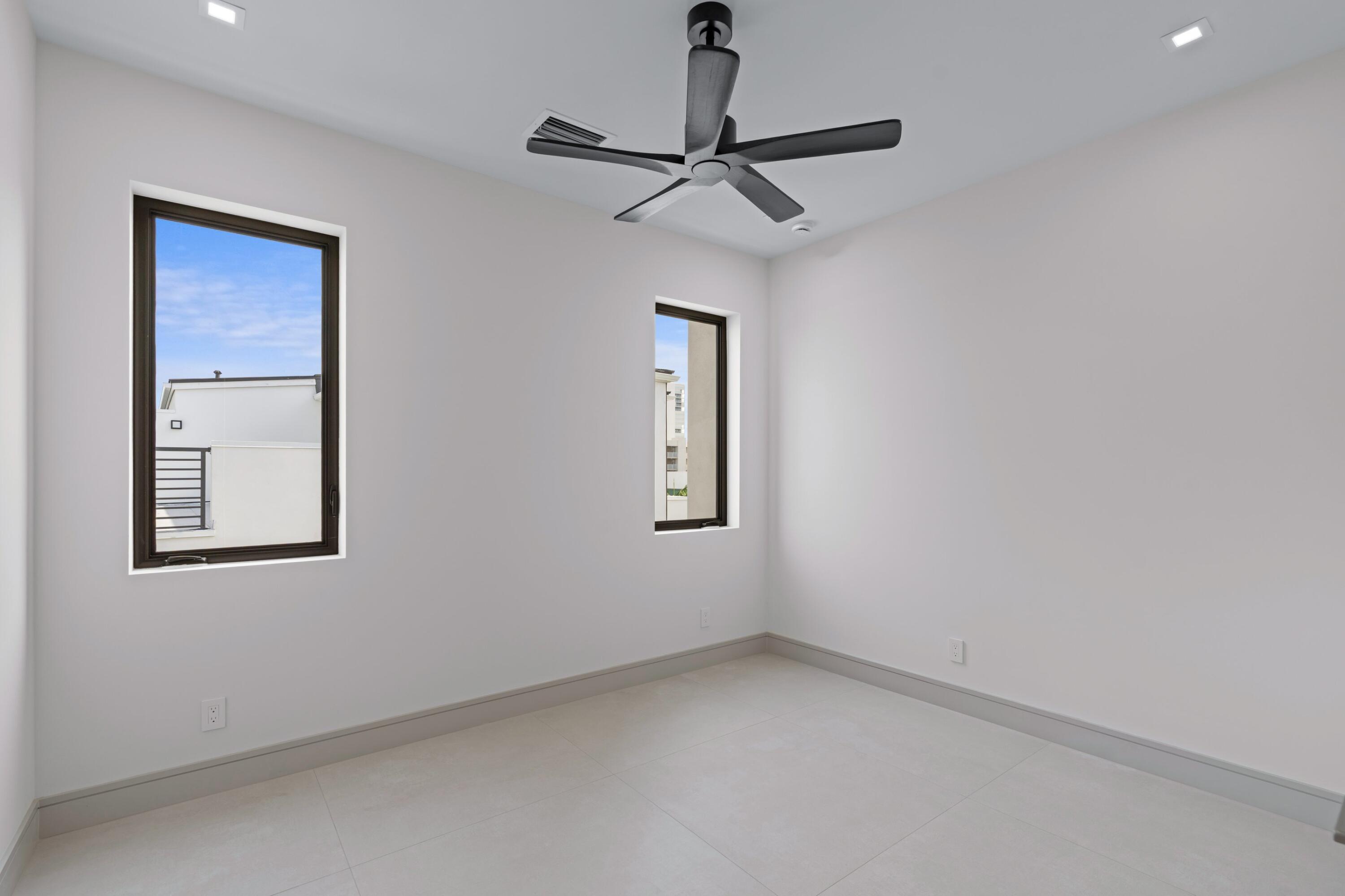 Empty bedroom with two windows, pale tile flooring, and a black ceiling fan.