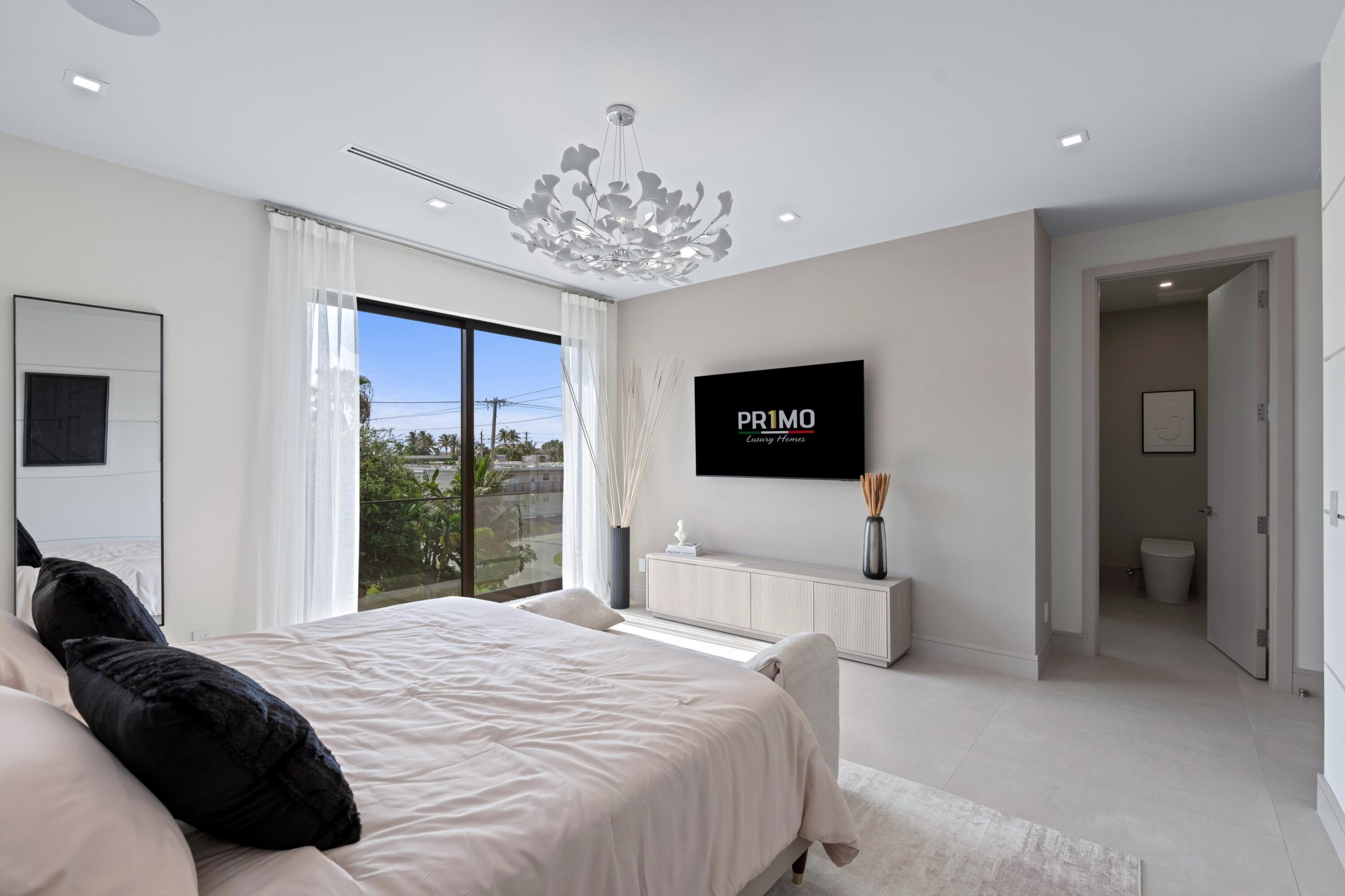 Bright staged bedroom with a large window, sculptural chandelier, and ensuite bathroom doorway.