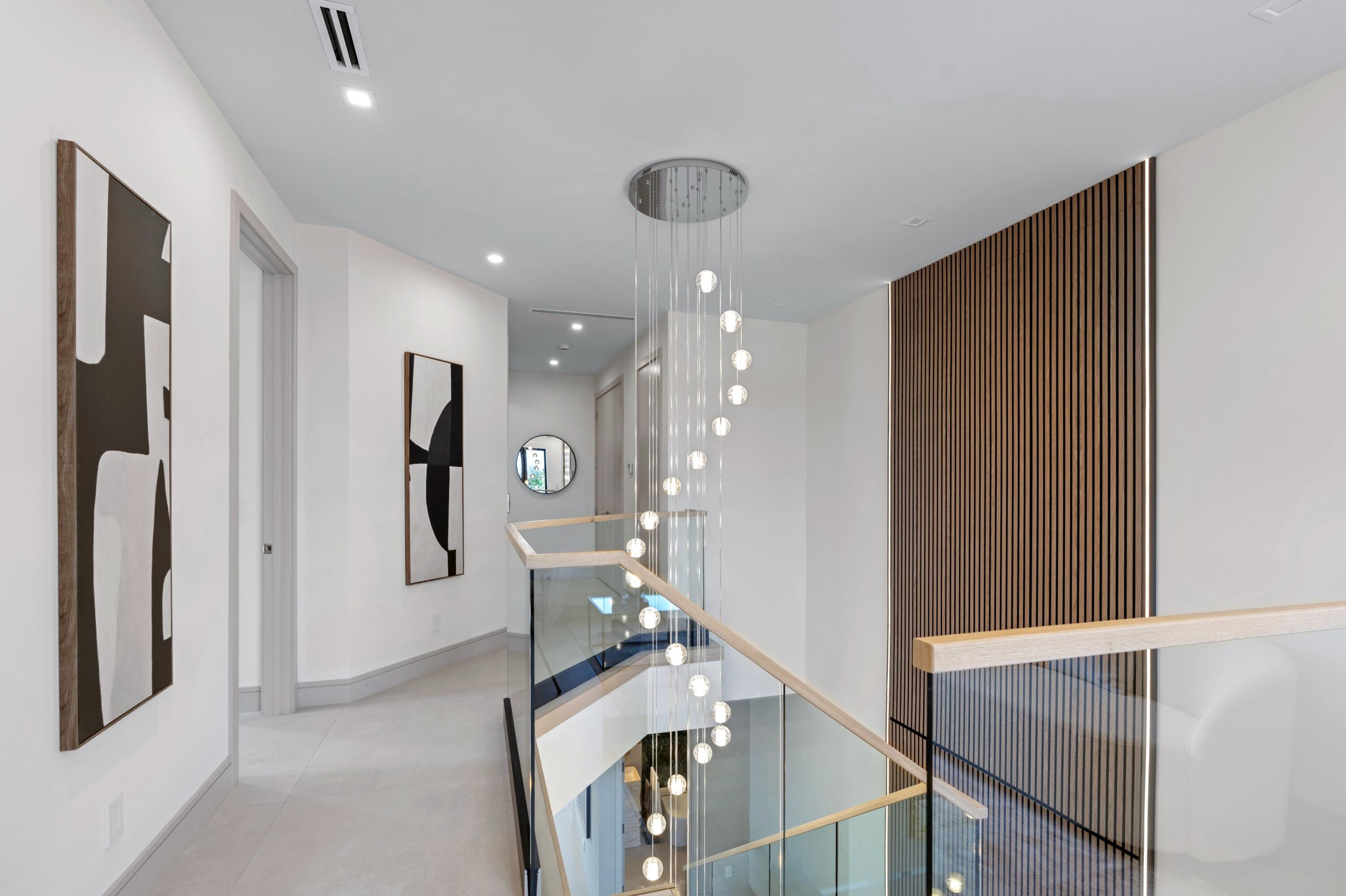 Upstairs hallway overlooking a staircase, with a glass railing, sculptural pendant lights, and a slatted wood accent wall.