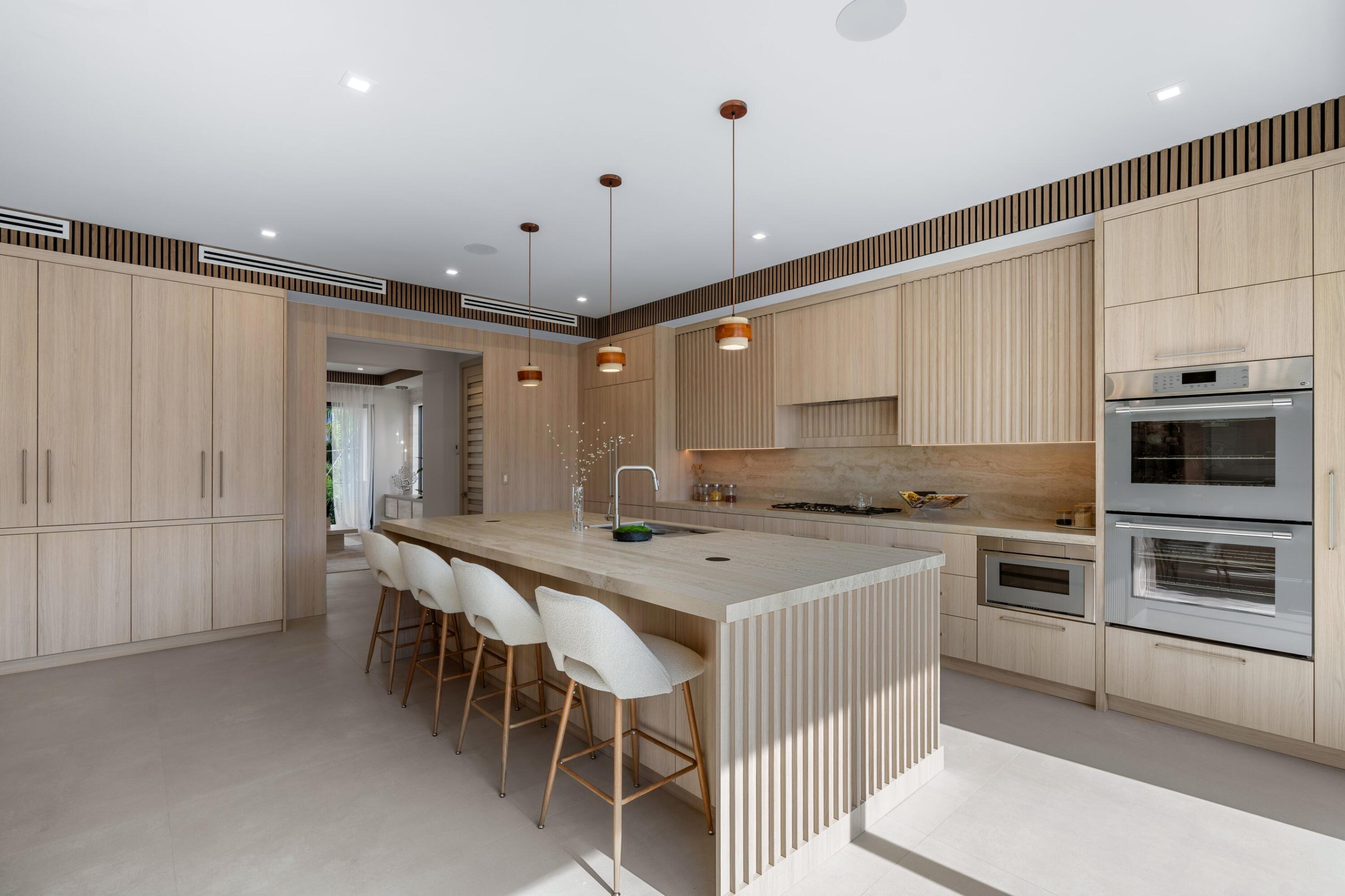 Modern kitchen with a large island, pale wood cabinetry, white stools, and double wall ovens.