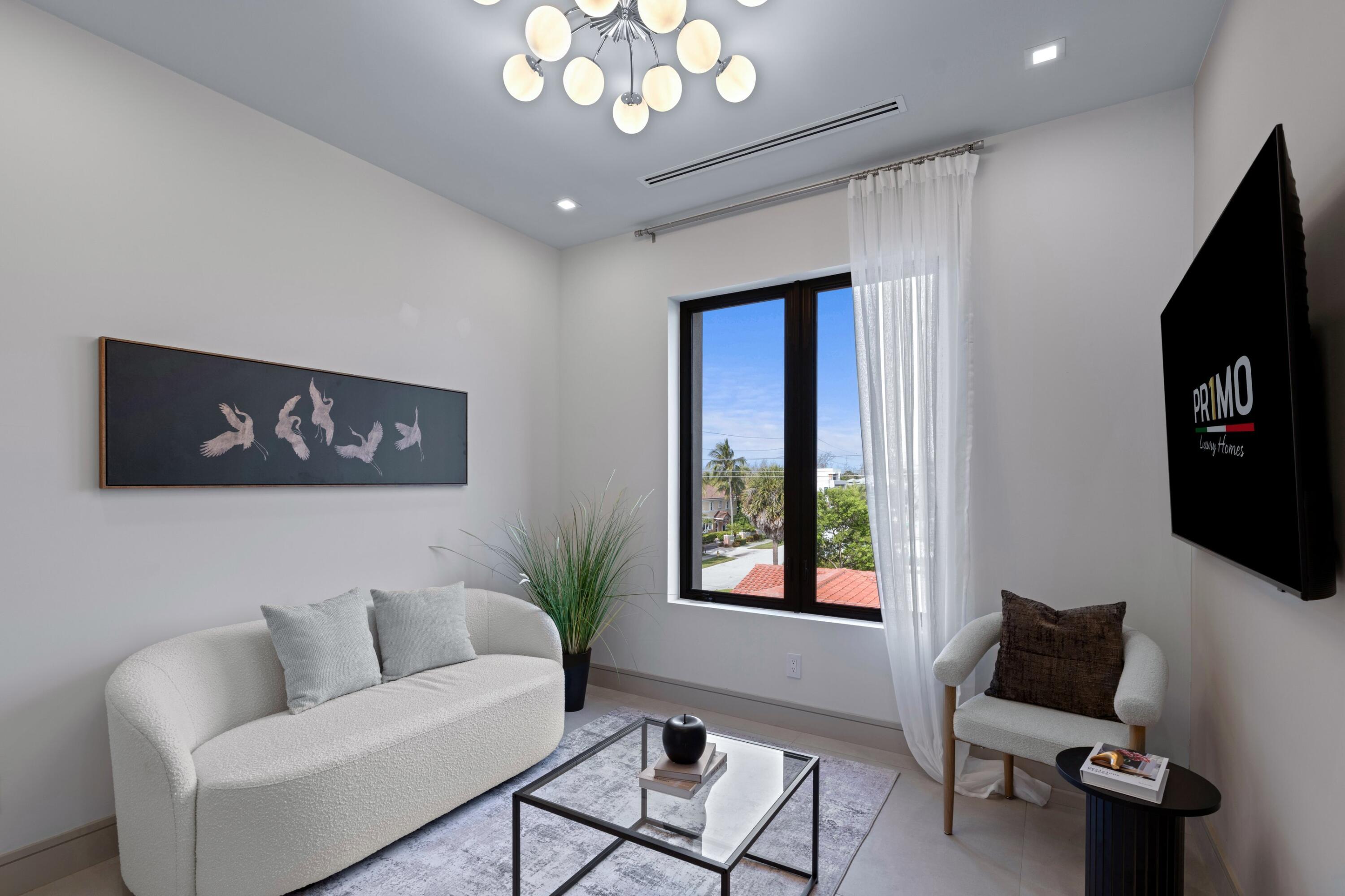 Living room with curved white sofa, glass coffee table, and large window overlooking palm trees.