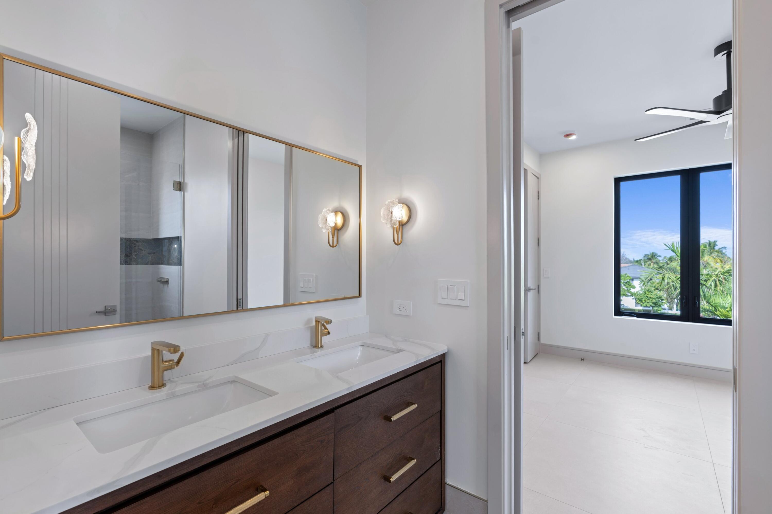 Modern bathroom with a double vanity, gold fixtures, and a doorway to a bright room.