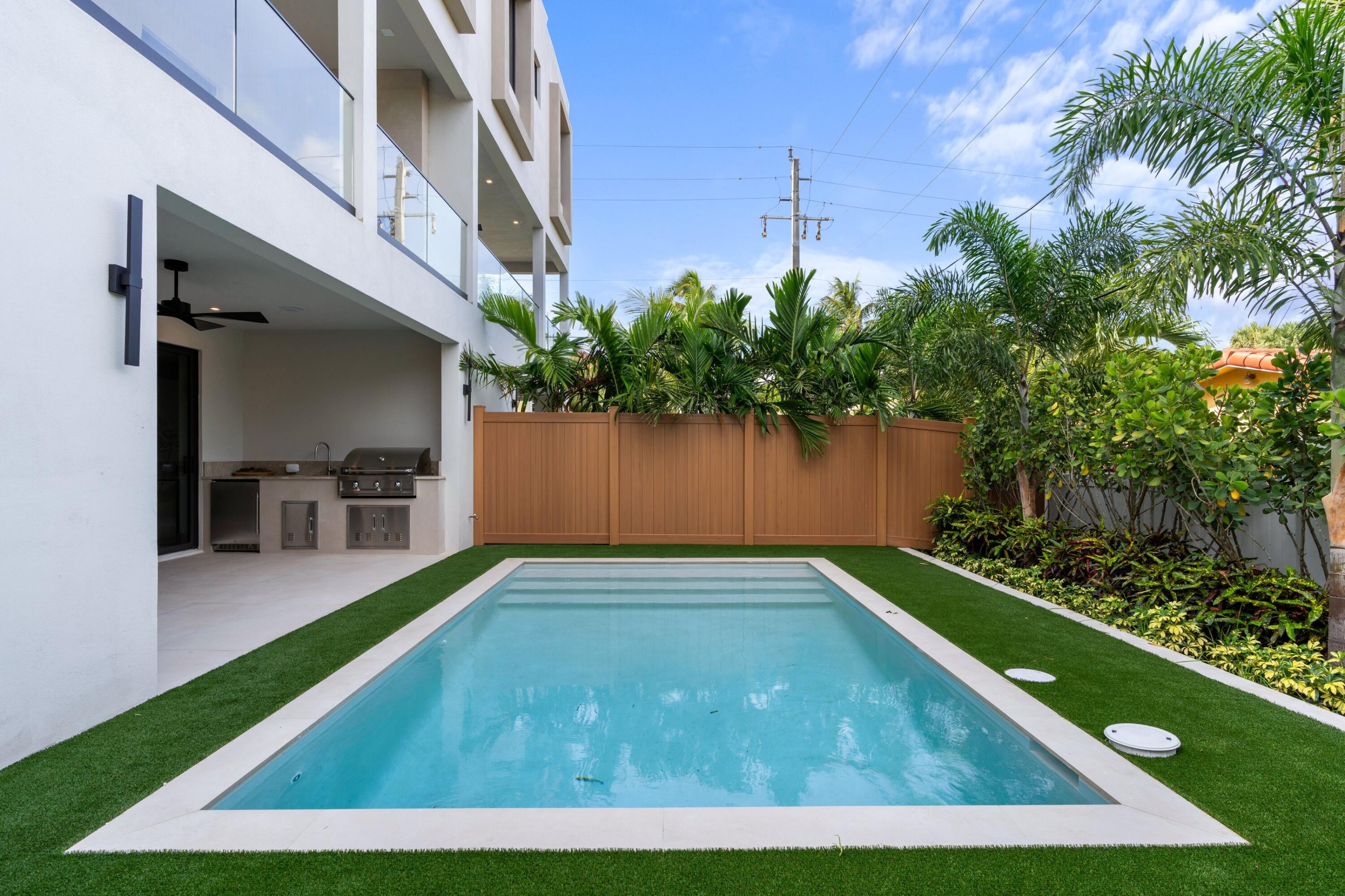 Rectangular backyard pool beside a modern white home, with covered outdoor kitchen and tropical landscaping.