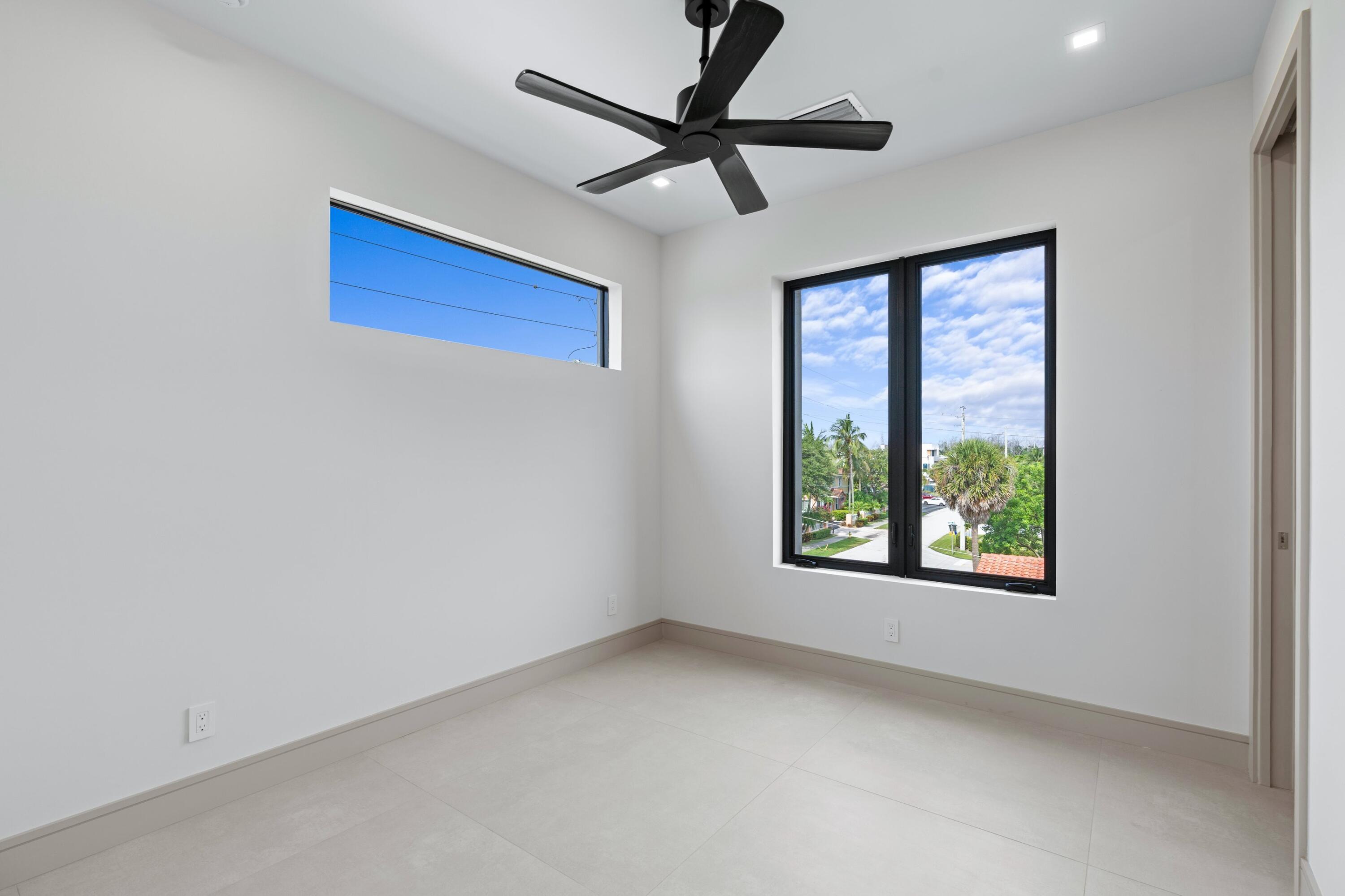 Empty room with pale tile floors, a black ceiling fan, and street views through wide windows.