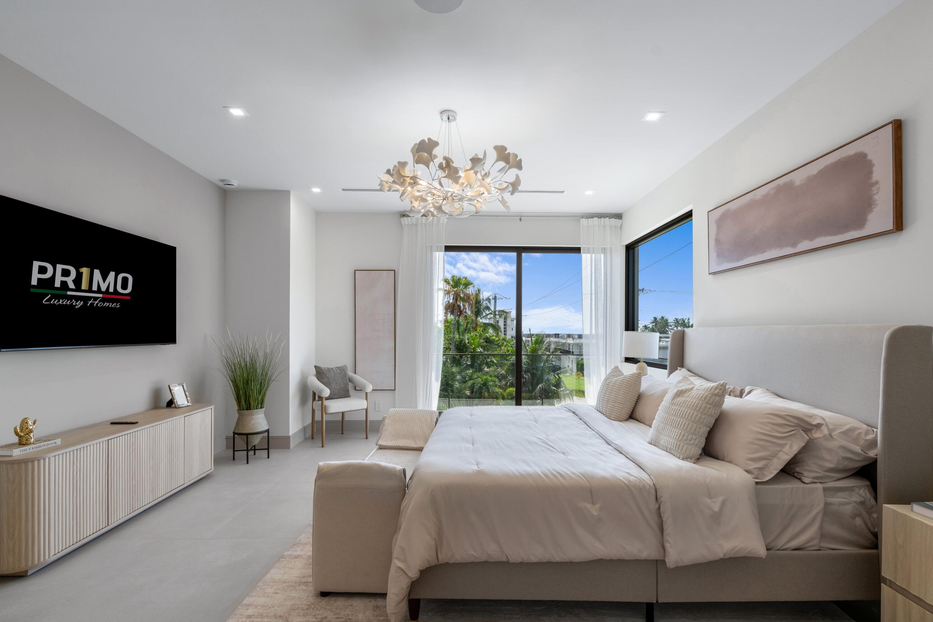 Bright staged bedroom with a large bed, sculptural chandelier, and windows overlooking palm trees.