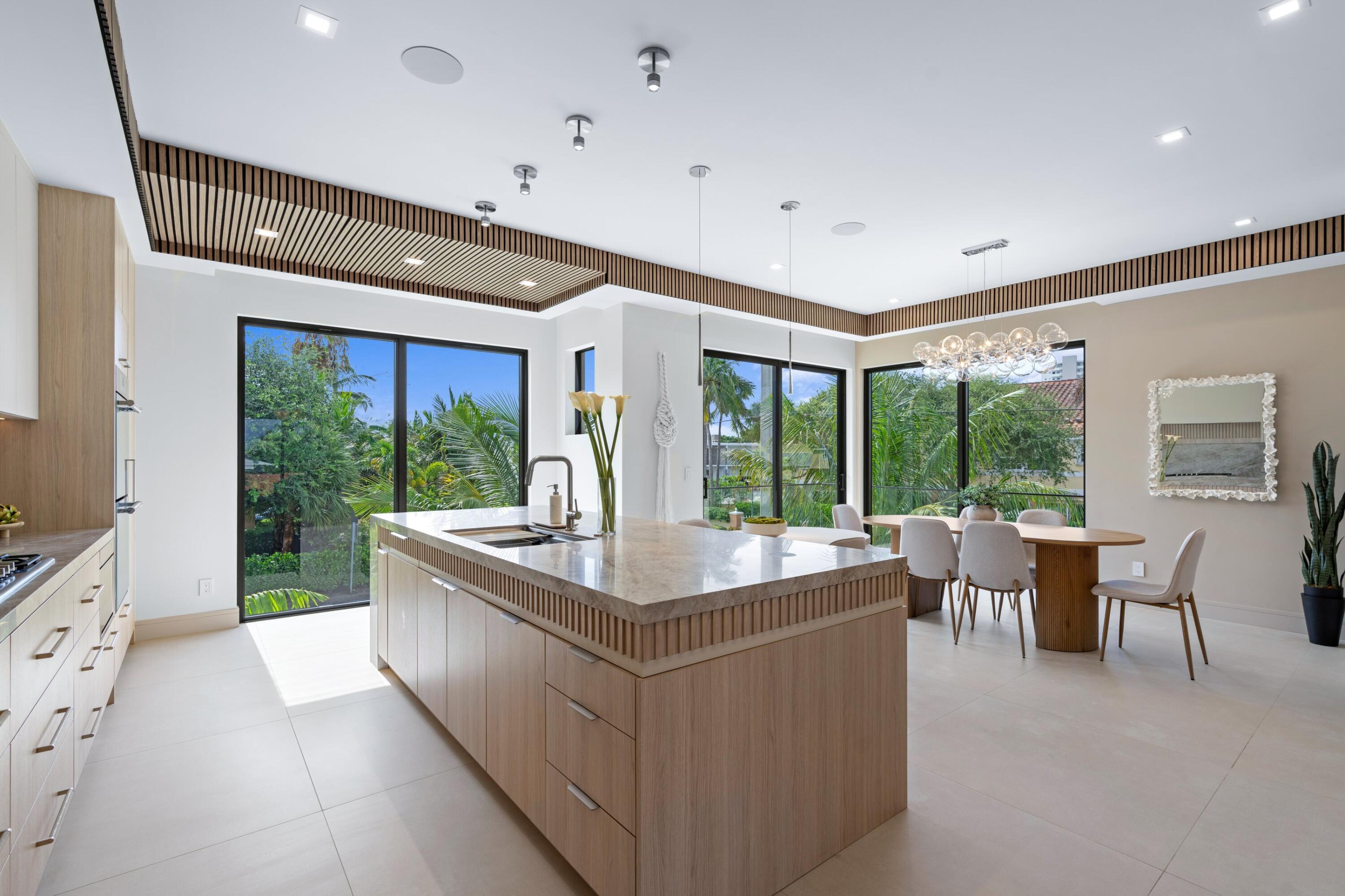 Modern kitchen with a large wood island, opening to a dining area and tropical greenery.