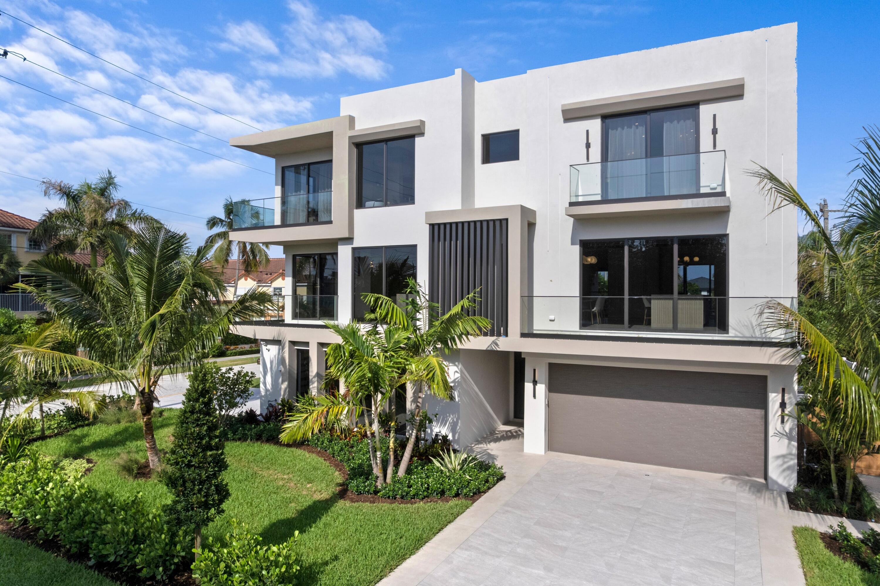 Modern white home with glass balconies, tropical landscaping, and a wide driveway.