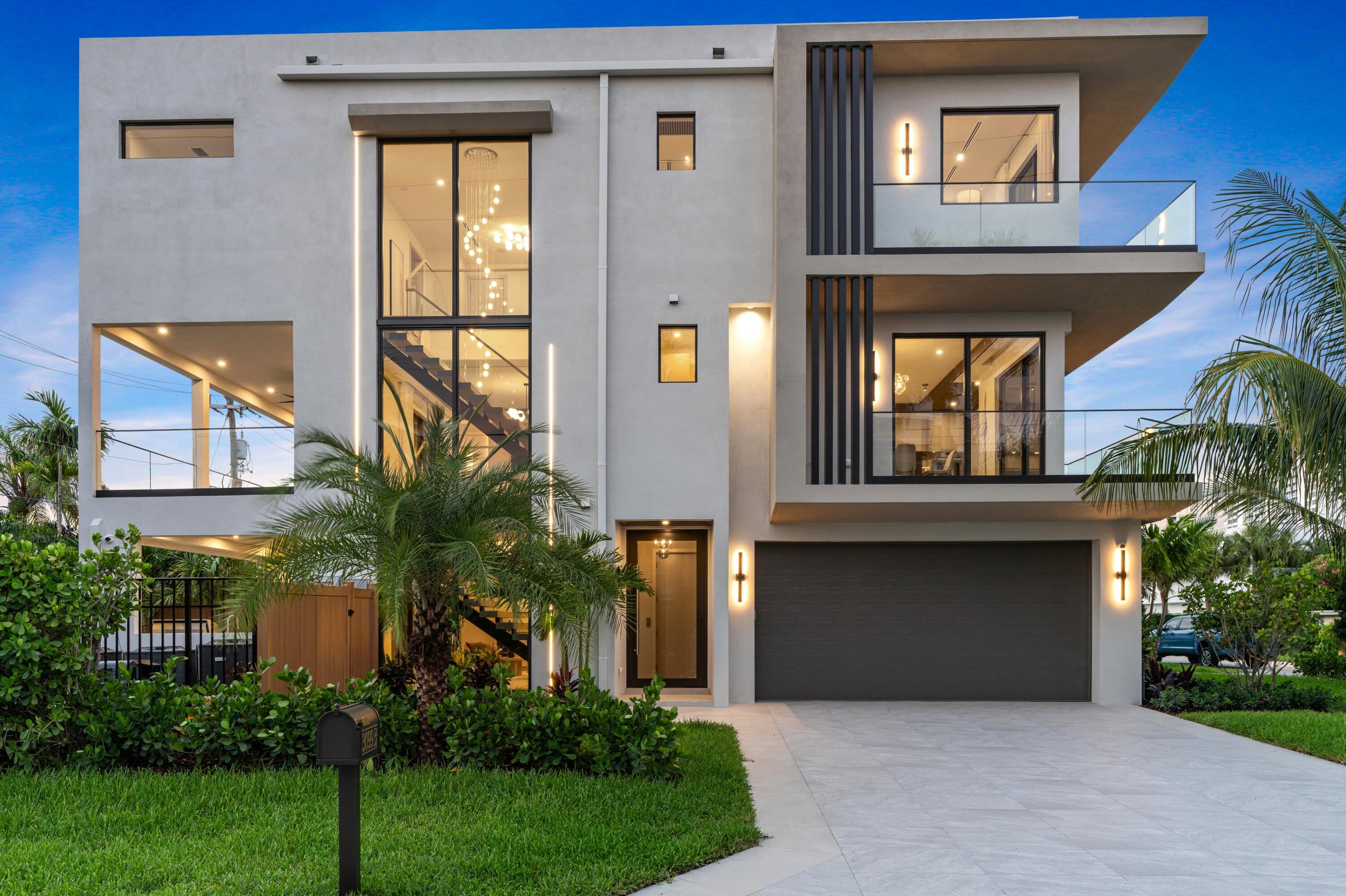 Modern three-story home with glass balconies, illuminated windows, and tropical landscaping at dusk.