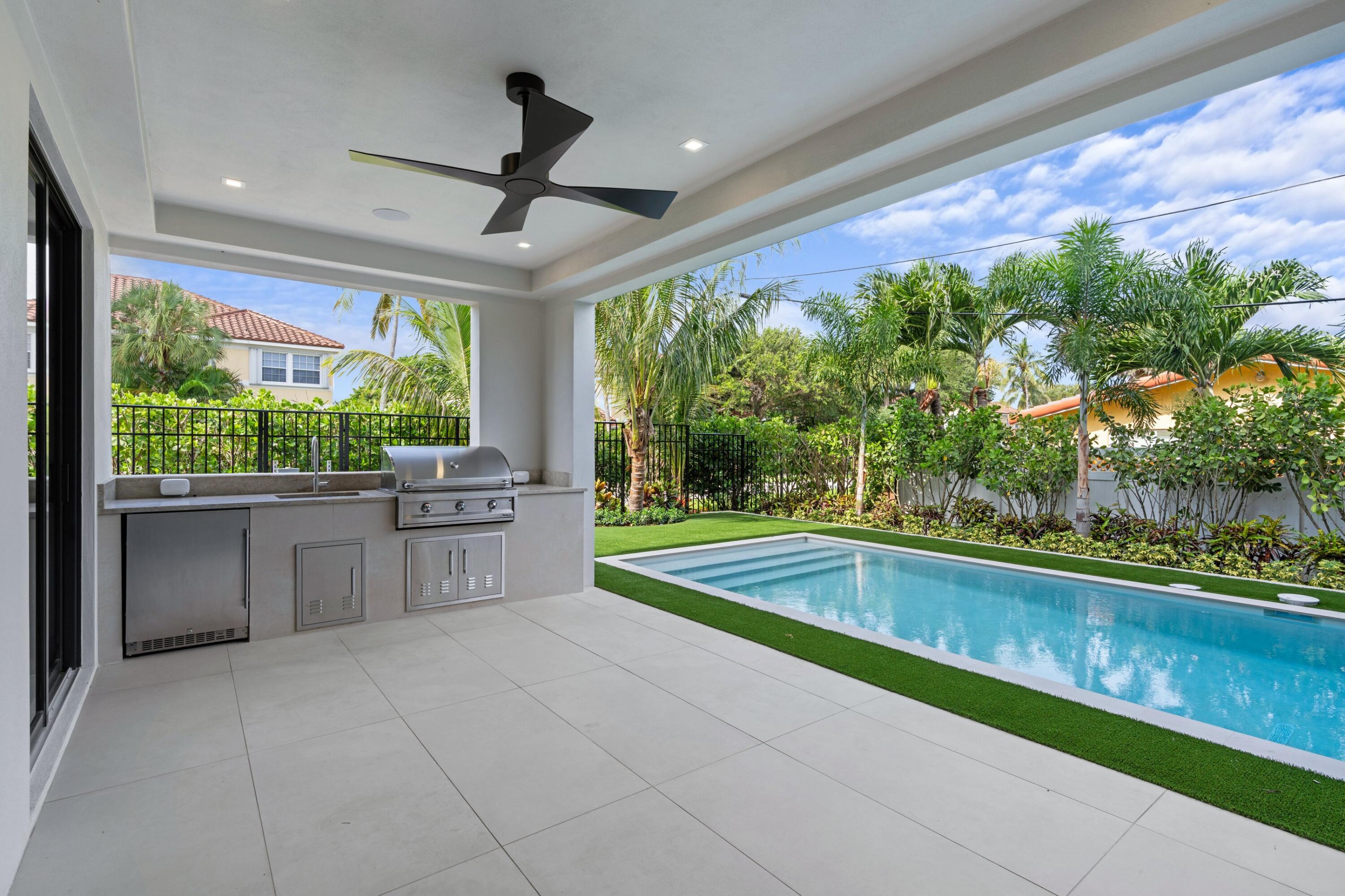 Covered patio with an outdoor kitchen overlooking a narrow swimming pool and palm-lined yard.