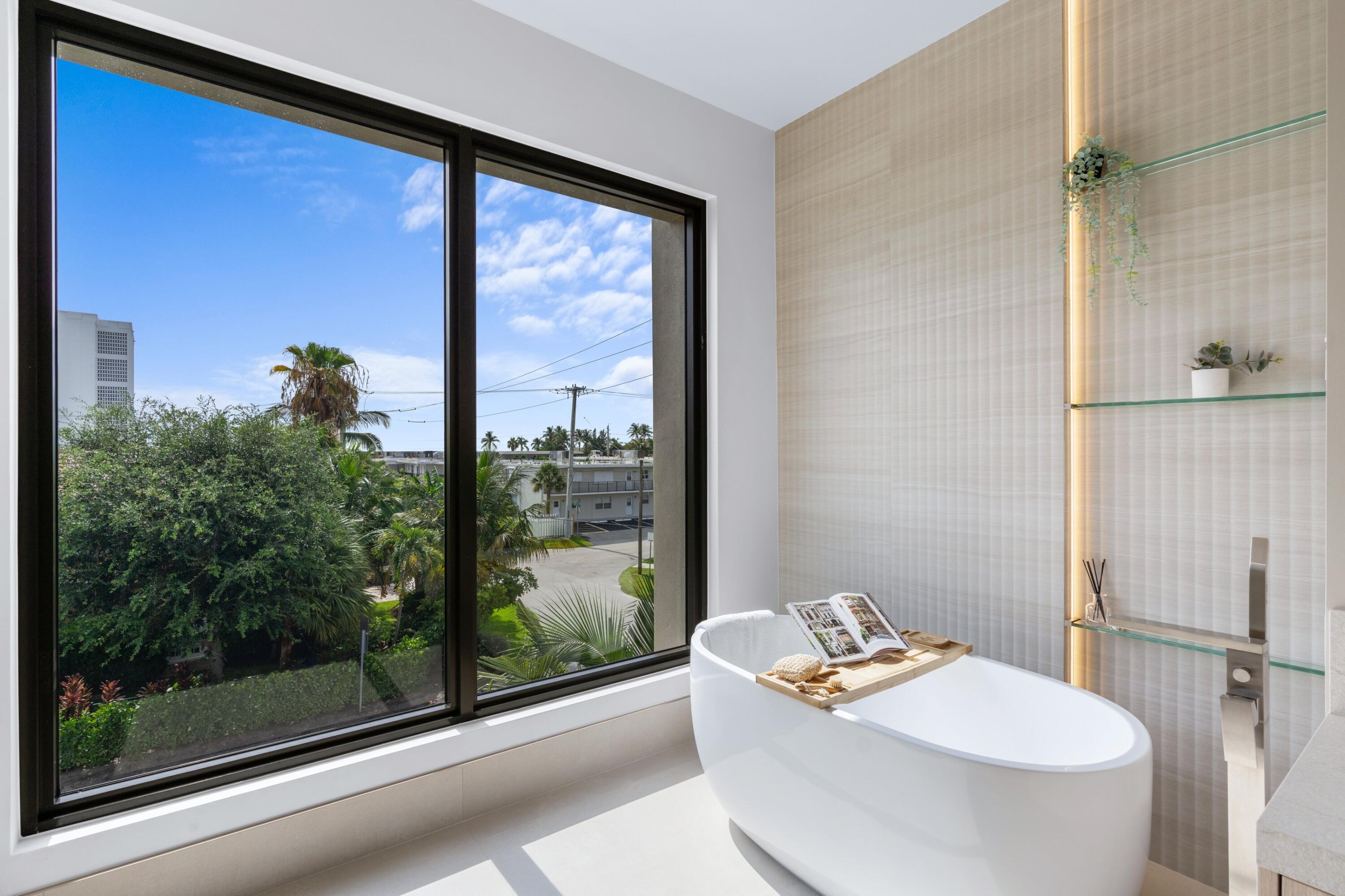 Bathroom with a freestanding tub beside a large window overlooking palm trees and nearby buildings.