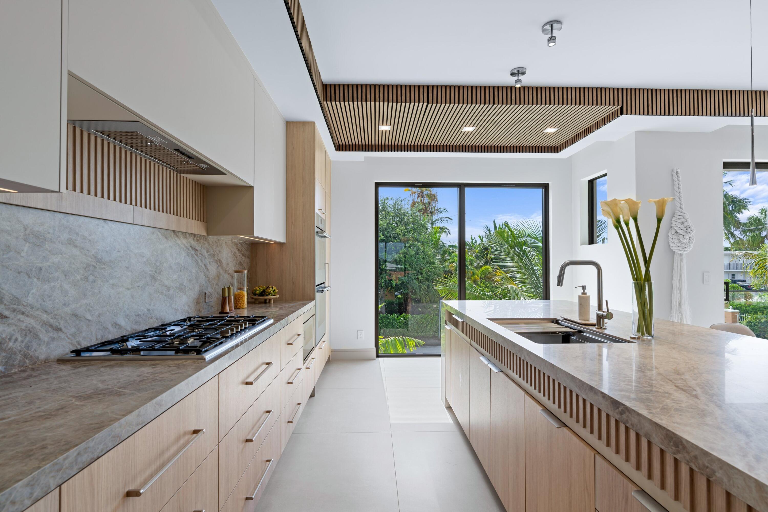 Modern kitchen with a stone island, wood cabinetry, and garden views through sliding glass doors.
