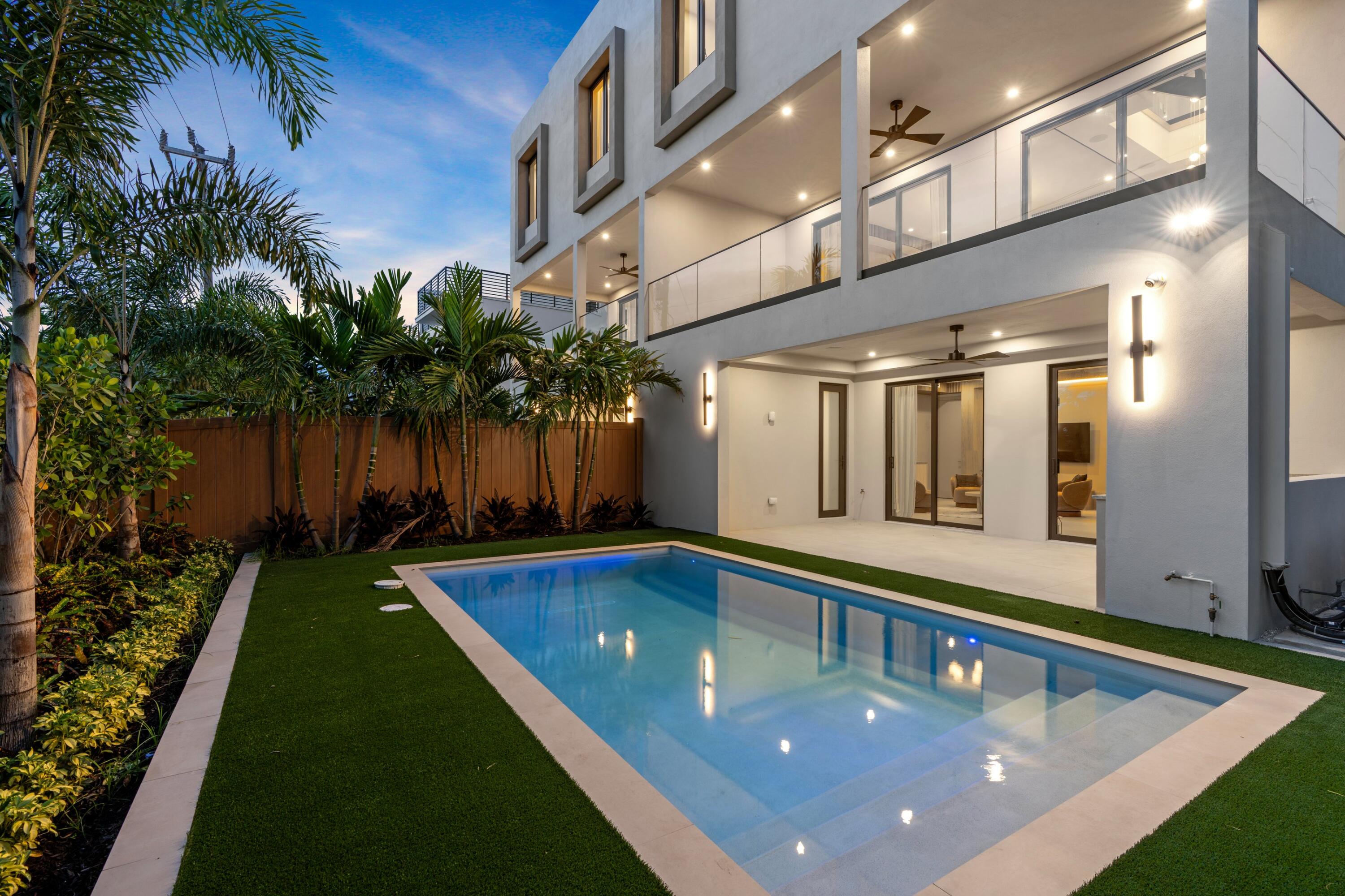 Modern home with a lit swimming pool, glass balconies, and tropical landscaping at dusk.