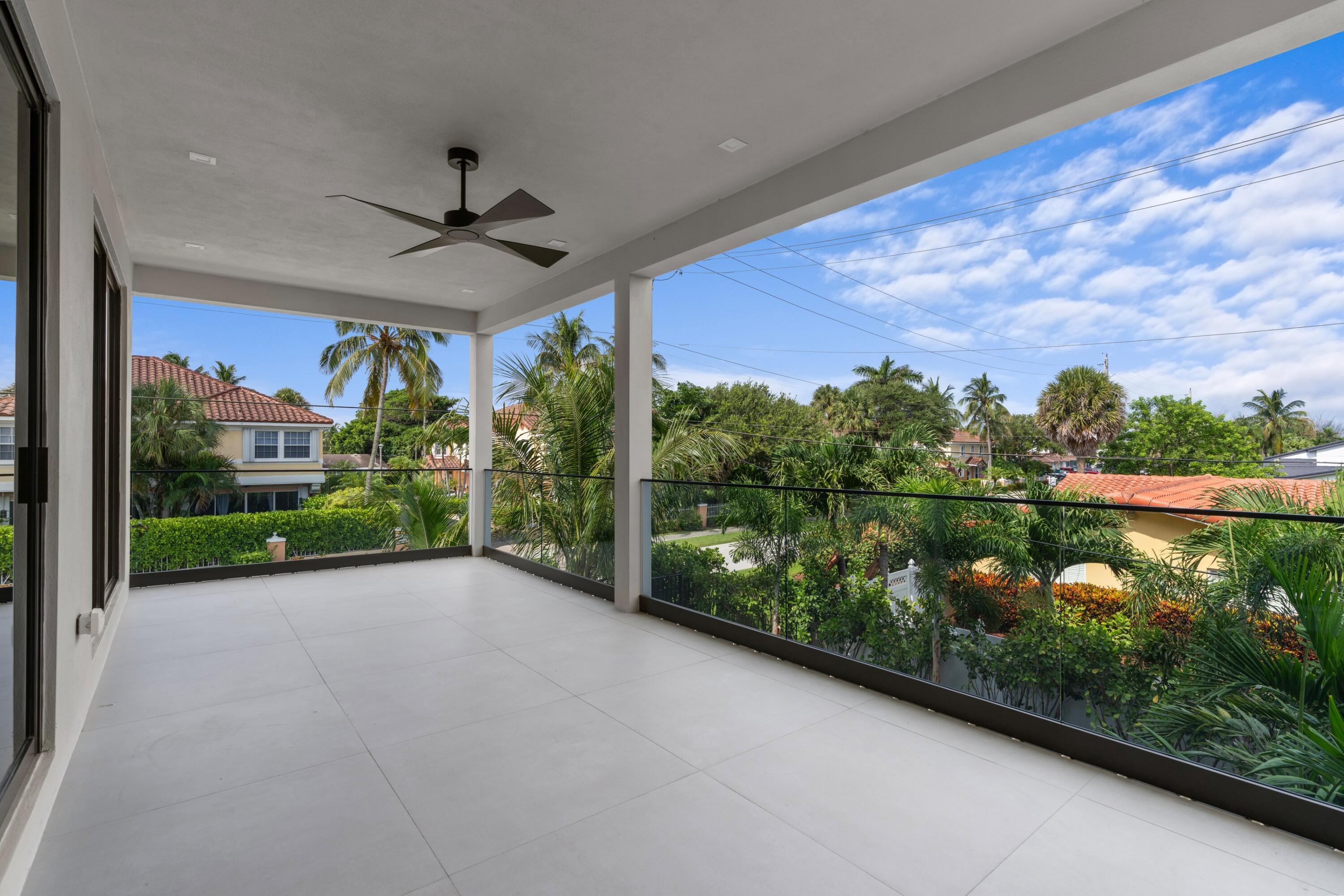 Covered tiled balcony with glass railings, ceiling fan, and palm-lined neighborhood views.