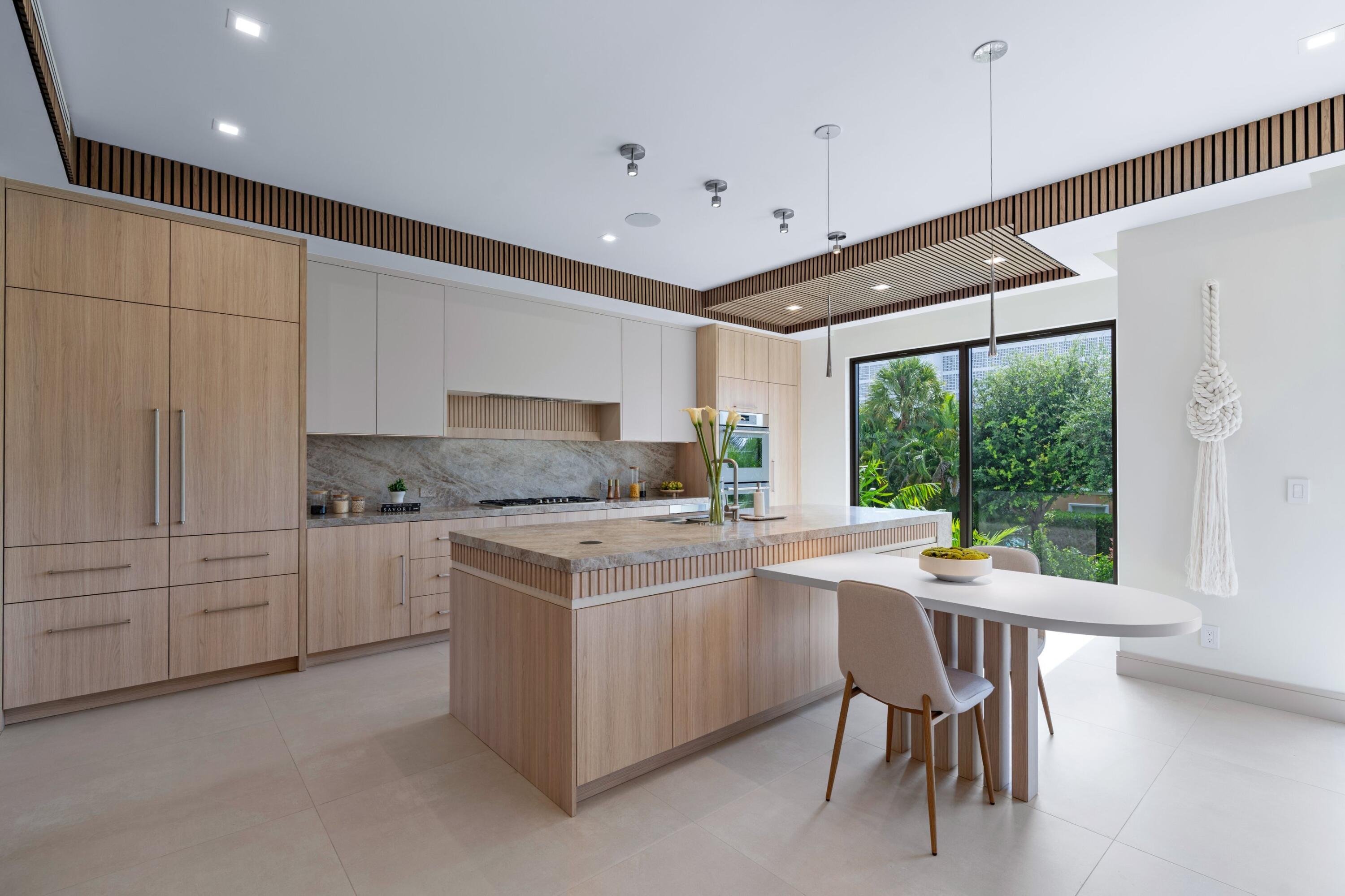 Modern kitchen with a stone island, pale wood cabinetry, and a dining table beside garden-facing windows.