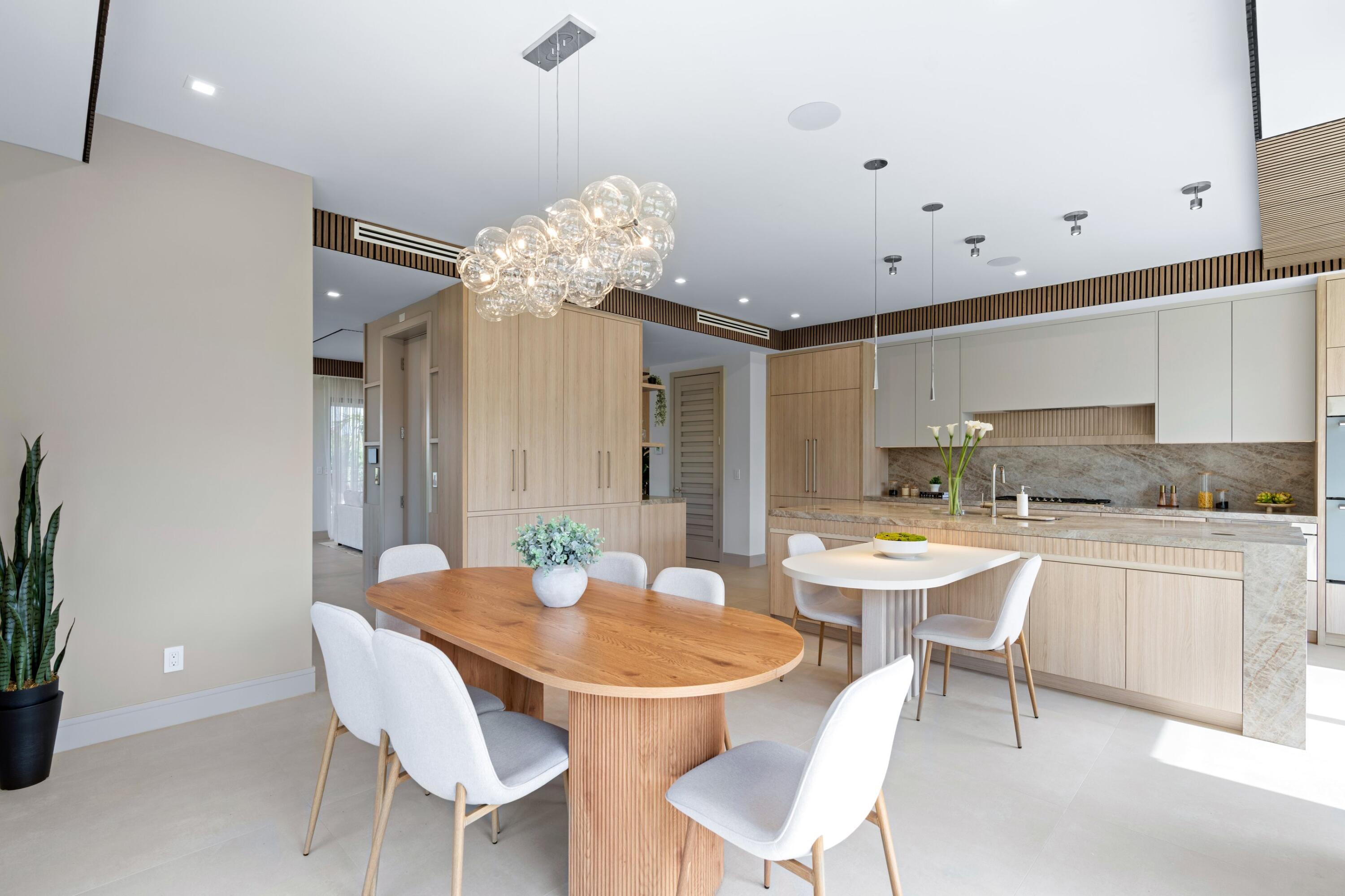 Modern kitchen and dining area with pale wood cabinetry, stone counters, and a sculptural glass chandelier.