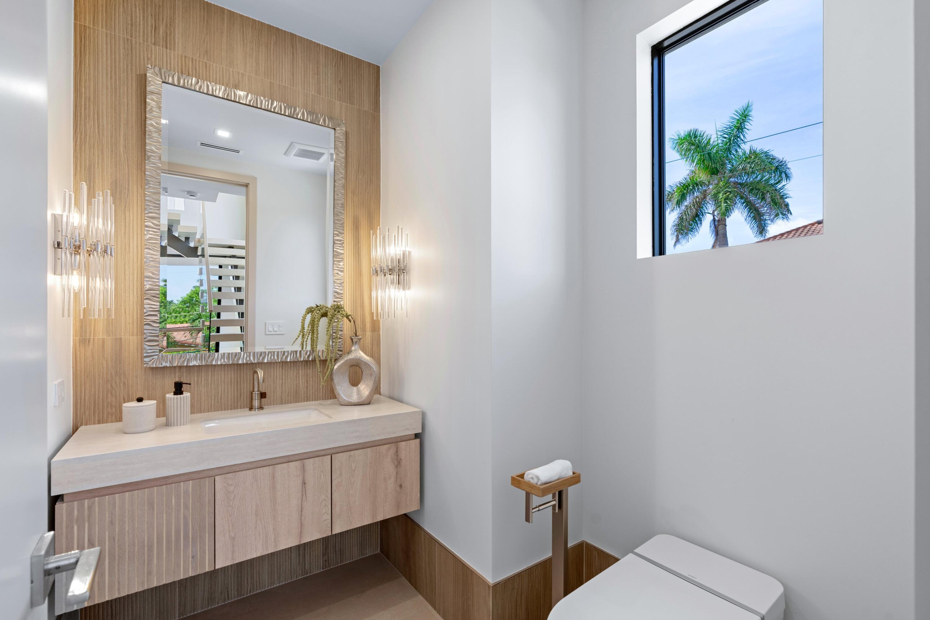 Modern bathroom with a floating wood vanity, sculptural mirror, and window overlooking a palm tree.