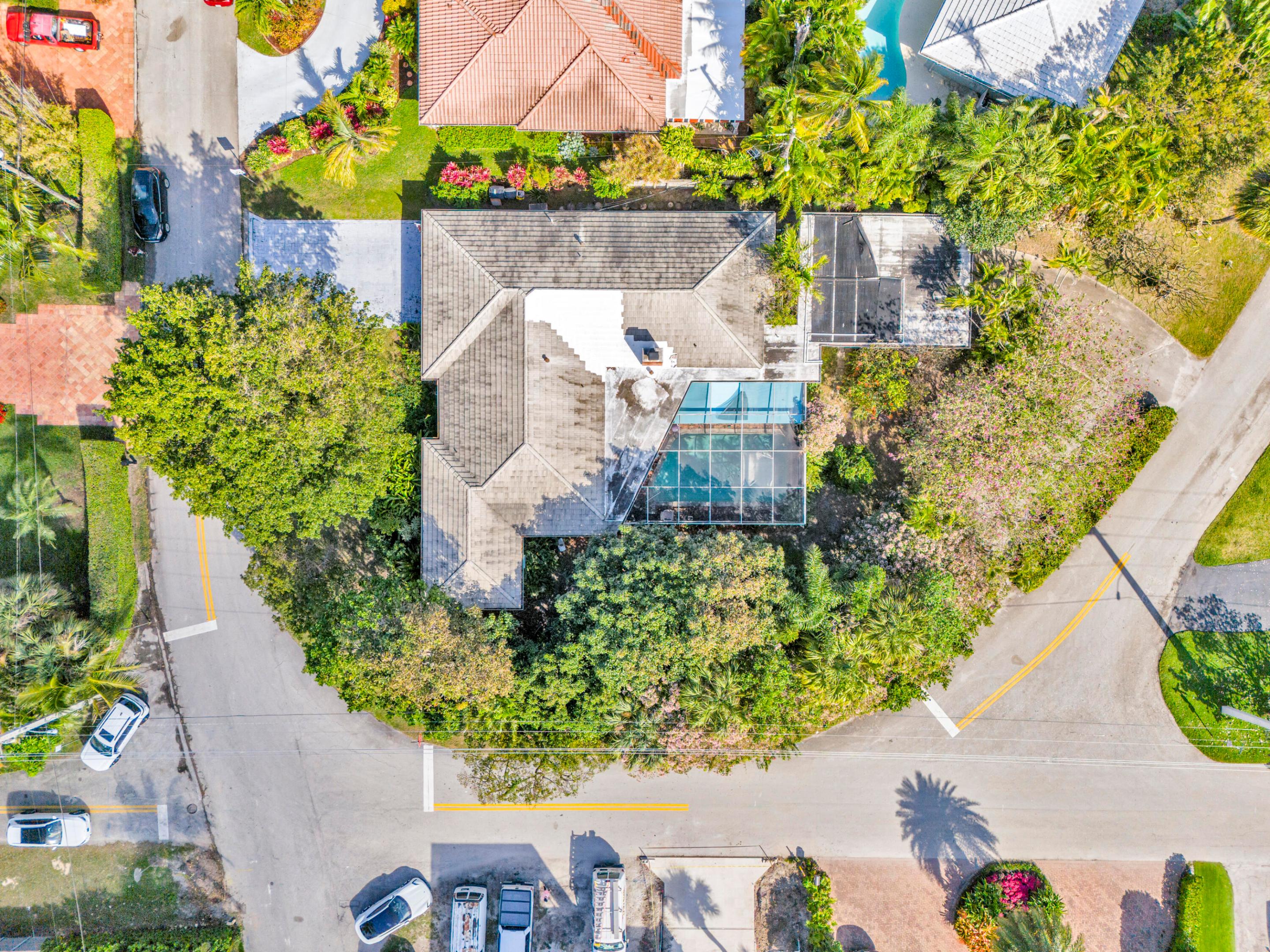 Aerial view of a landscaped home with a screened pool, surrounded by tropical trees at a street corner.