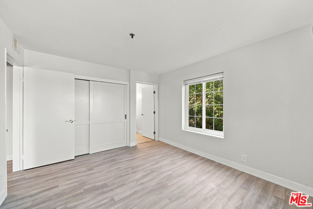 Empty bedroom with sliding closet doors, light wood-look flooring, and a window overlooking greenery.