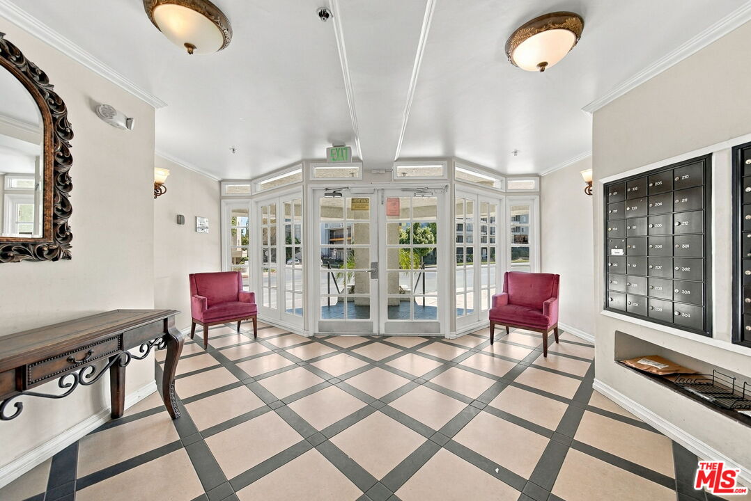 Apartment lobby with a glass entrance, wall of mailboxes, geometric tile floor, and burgundy chairs.