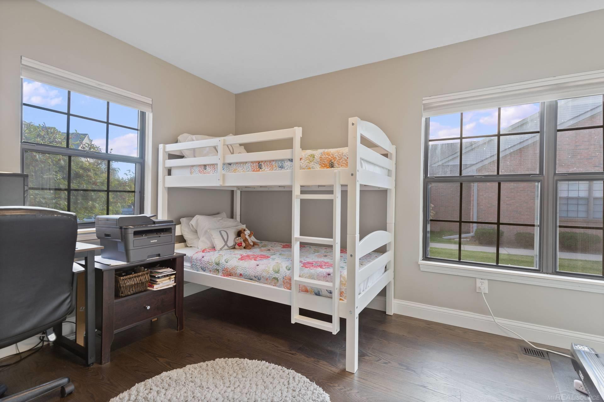 Bedroom with a white bunk bed, floral bedding, two large windows, and dark hardwood floors.