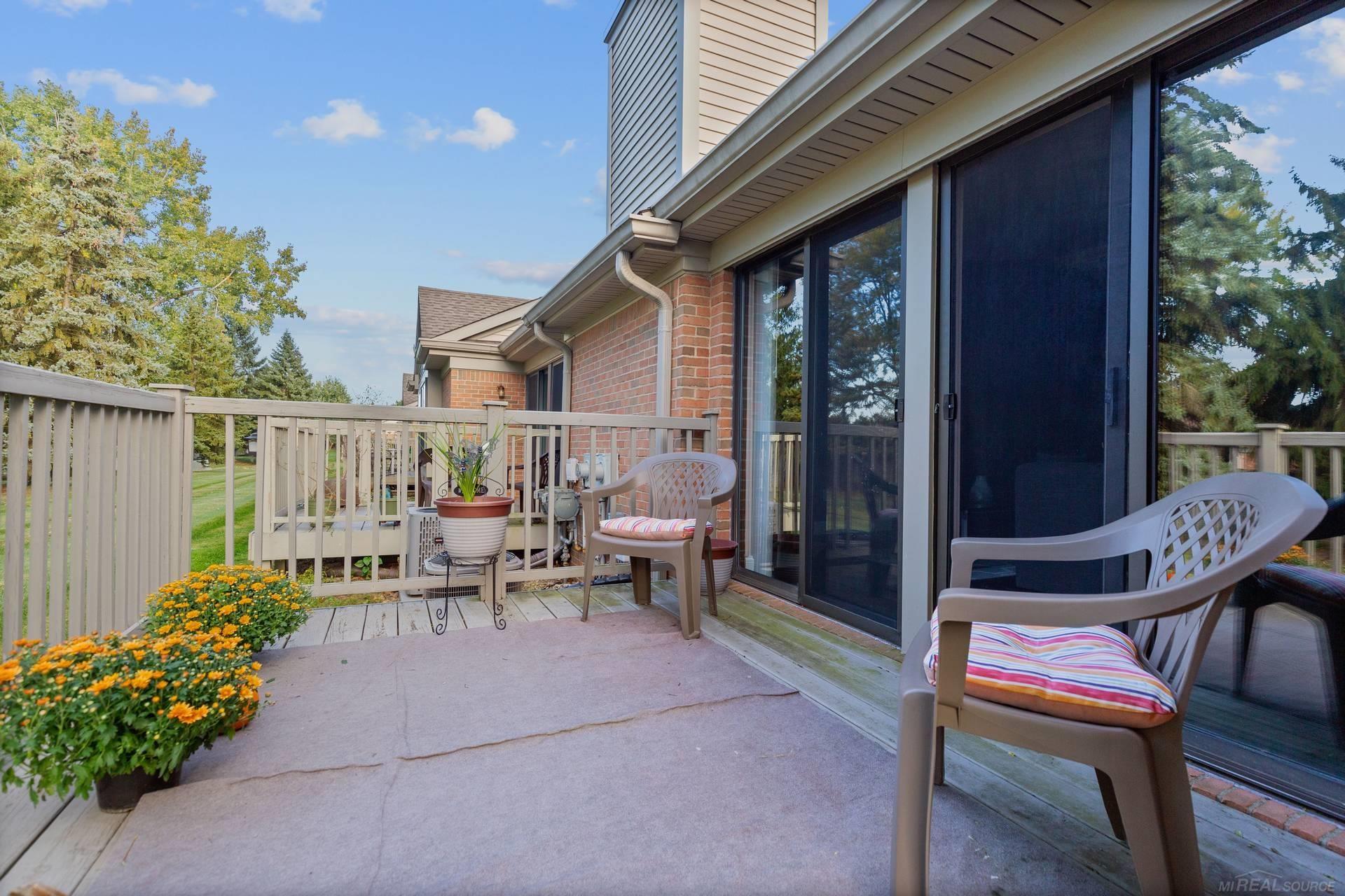 Furnished patio beside a brick home, with sliding doors, deck railing, chairs, and flower planters.