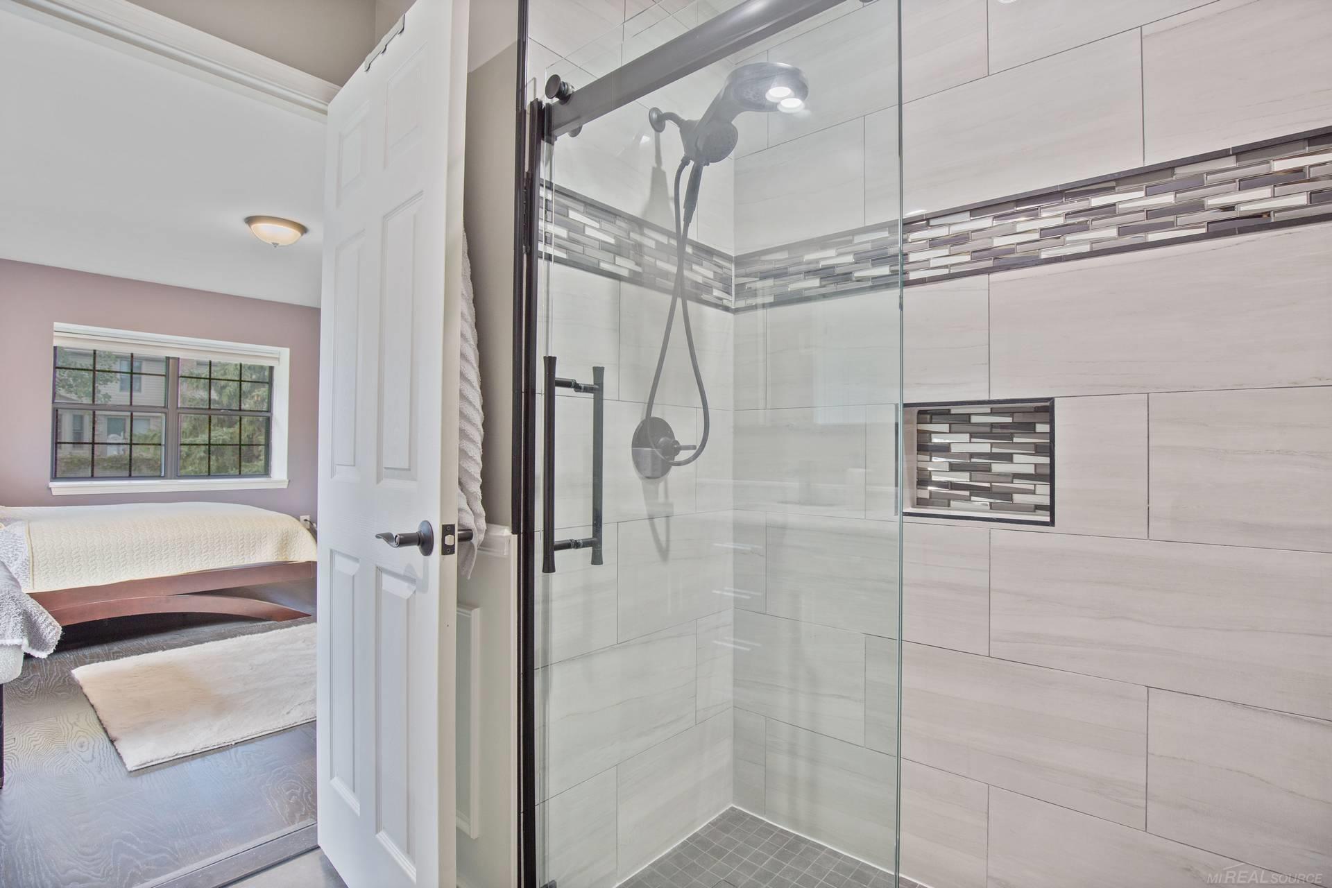 Walk-in shower with glass doors, light tile, and mosaic accents beside a bedroom.